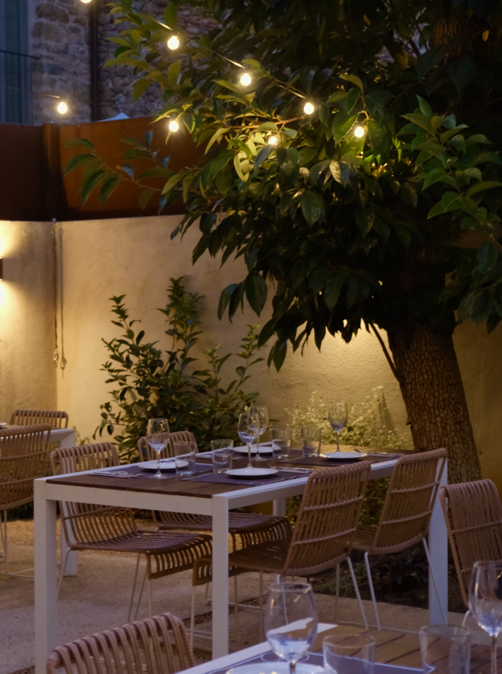 An outdoor dining area at dusk with a large tree wrapped in string lights, a white table set with wine glasses, plates, and utensils, and wicker chairs around the table.