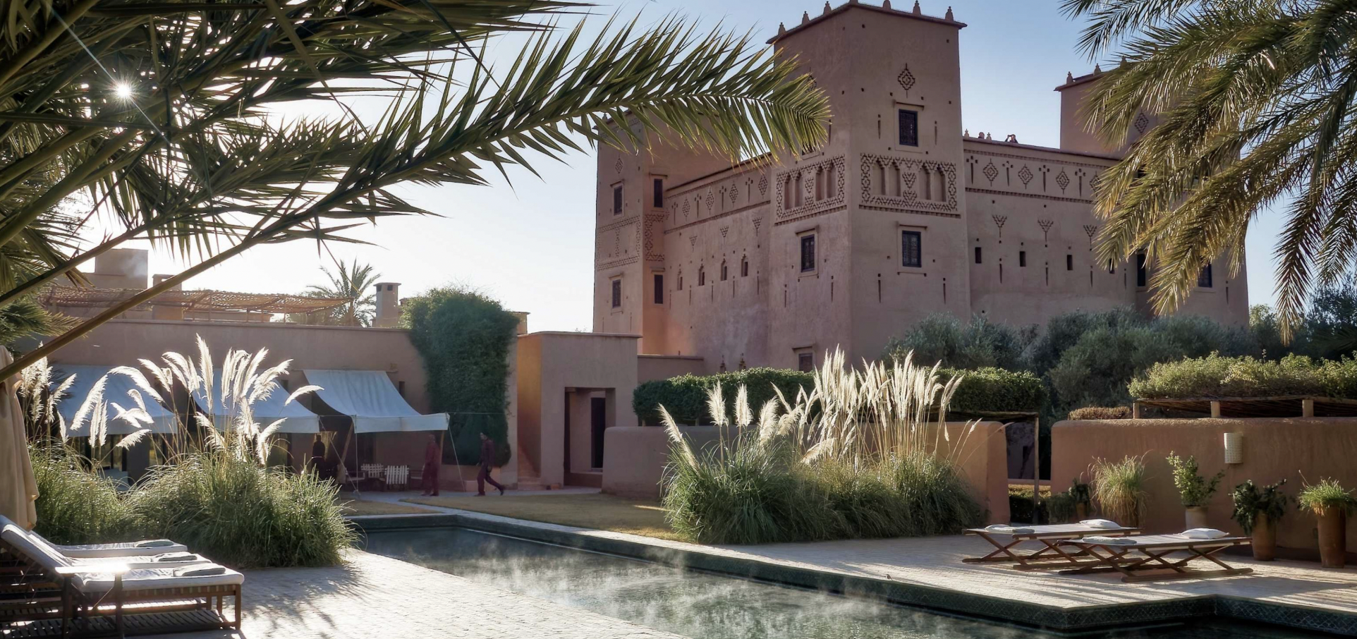 A castle-like building with desert-style architecture surrounded by lush desert plants, a pool, and lounge chairs in a sunny outdoor setting.