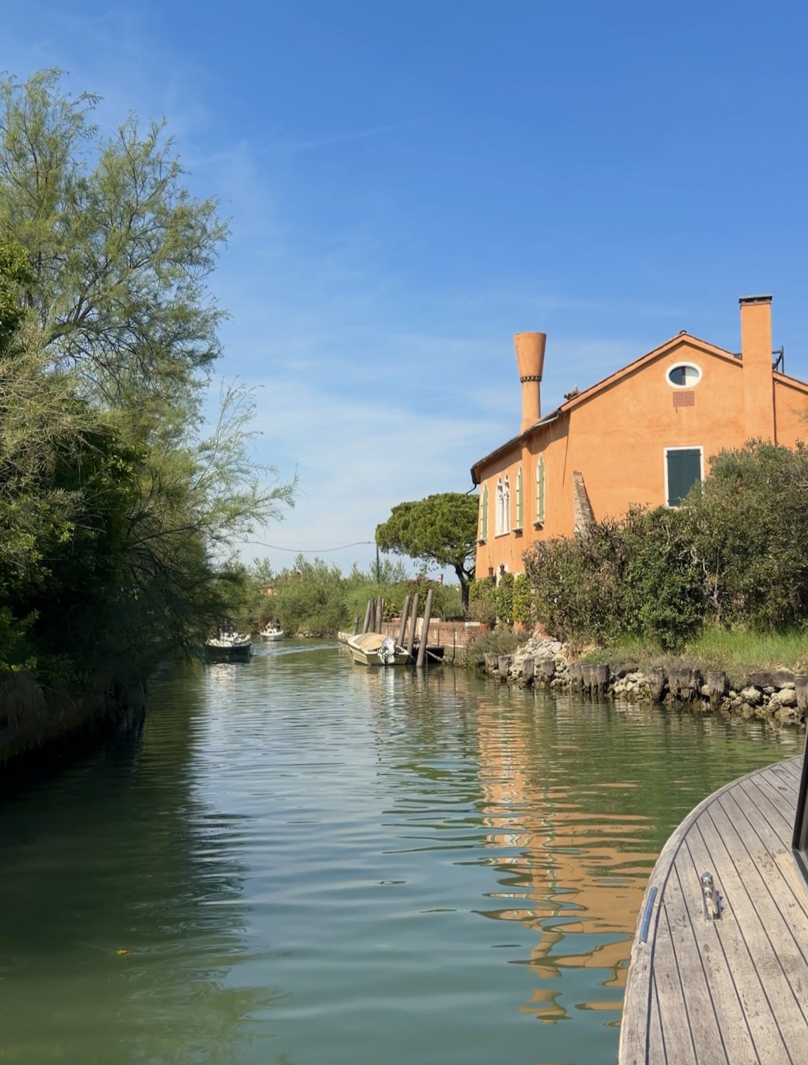 A canal with greenish water, lined with trees and plants, featuring a peach-colored house with chimneys on the right side and several boats docked along the water's edge. The sky is clear and blue.