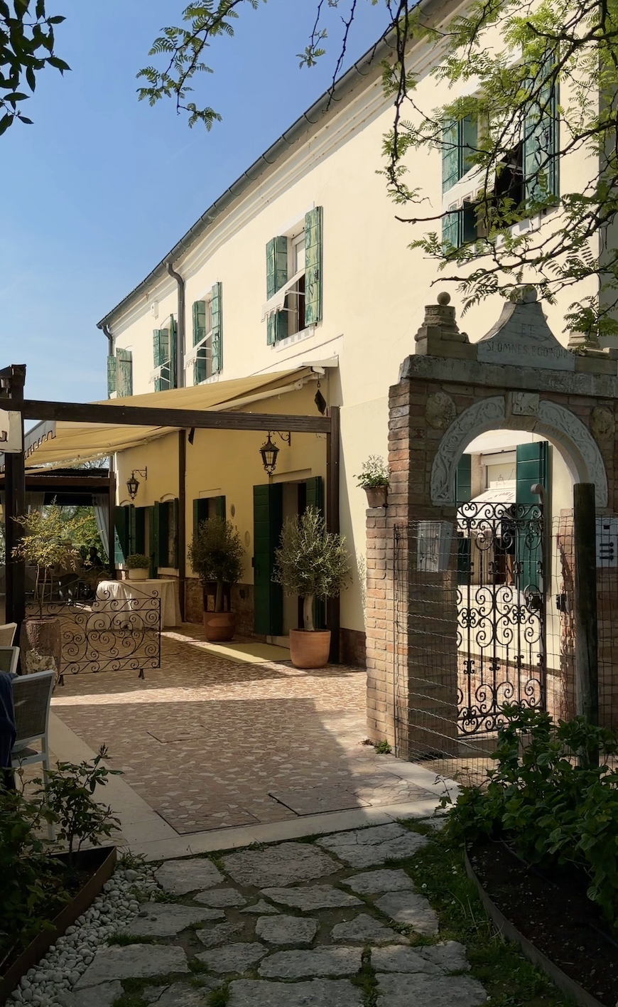 A charming outdoor courtyard with stone and brick paving, potted plants, and an entry gate with ornate ironwork. The building has green shutters, beige walls, and outdoor lanterns, with a yellow awning providing shade.