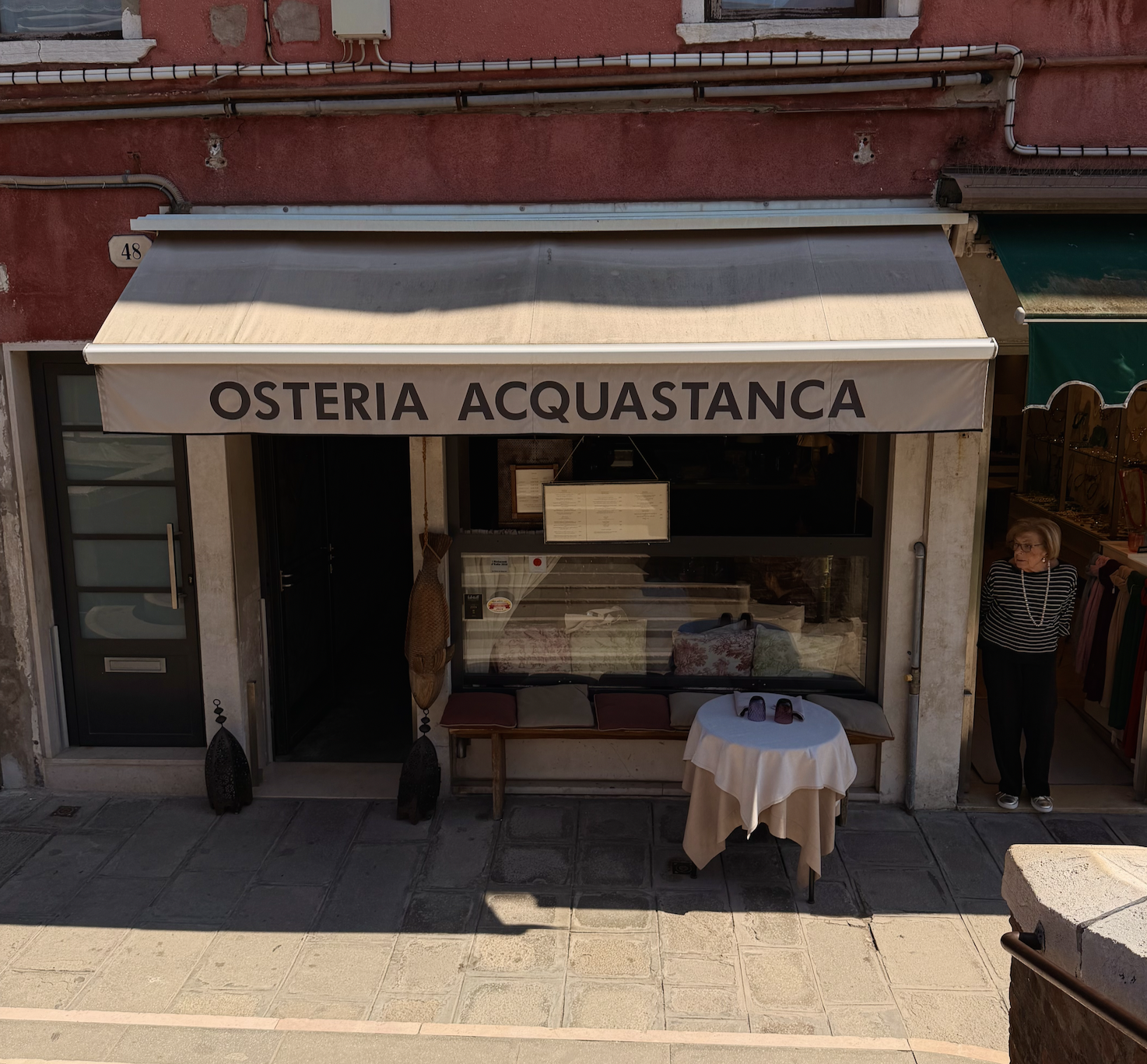 The storefront of Osteria Acquastanca, a restaurant, with a beige awning displaying its name. There is a table with shoes and a tablecloth outside, and an older woman standing near clothing on the right. The building has a red exterior with visible pipes and a door on the left.