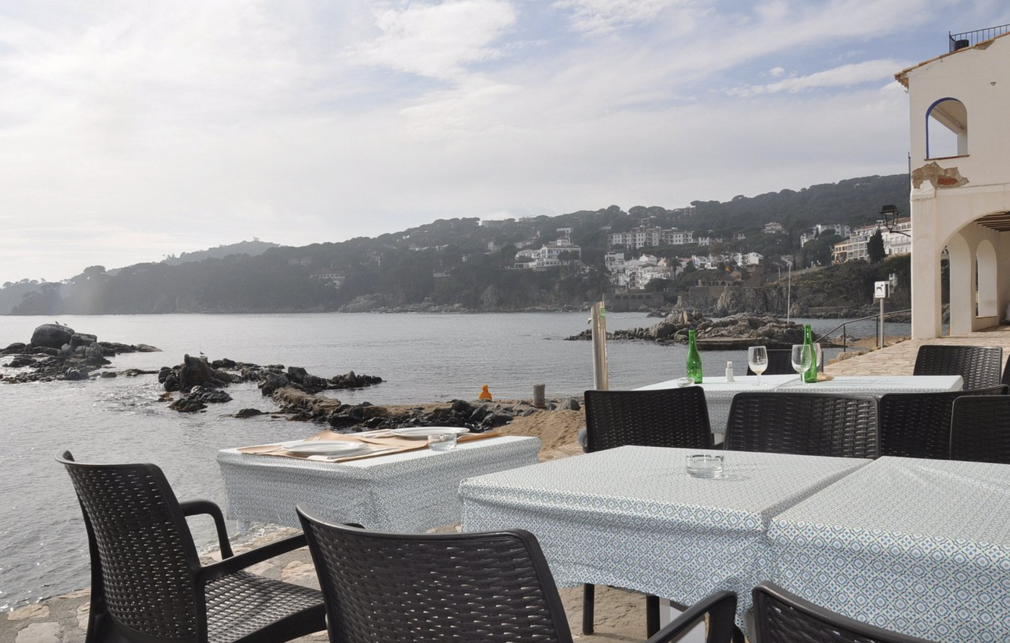 Outdoor dining tables with glass bottles and glasses, overlooking a rocky waterfront with a hillside town in the background on a partly cloudy day.