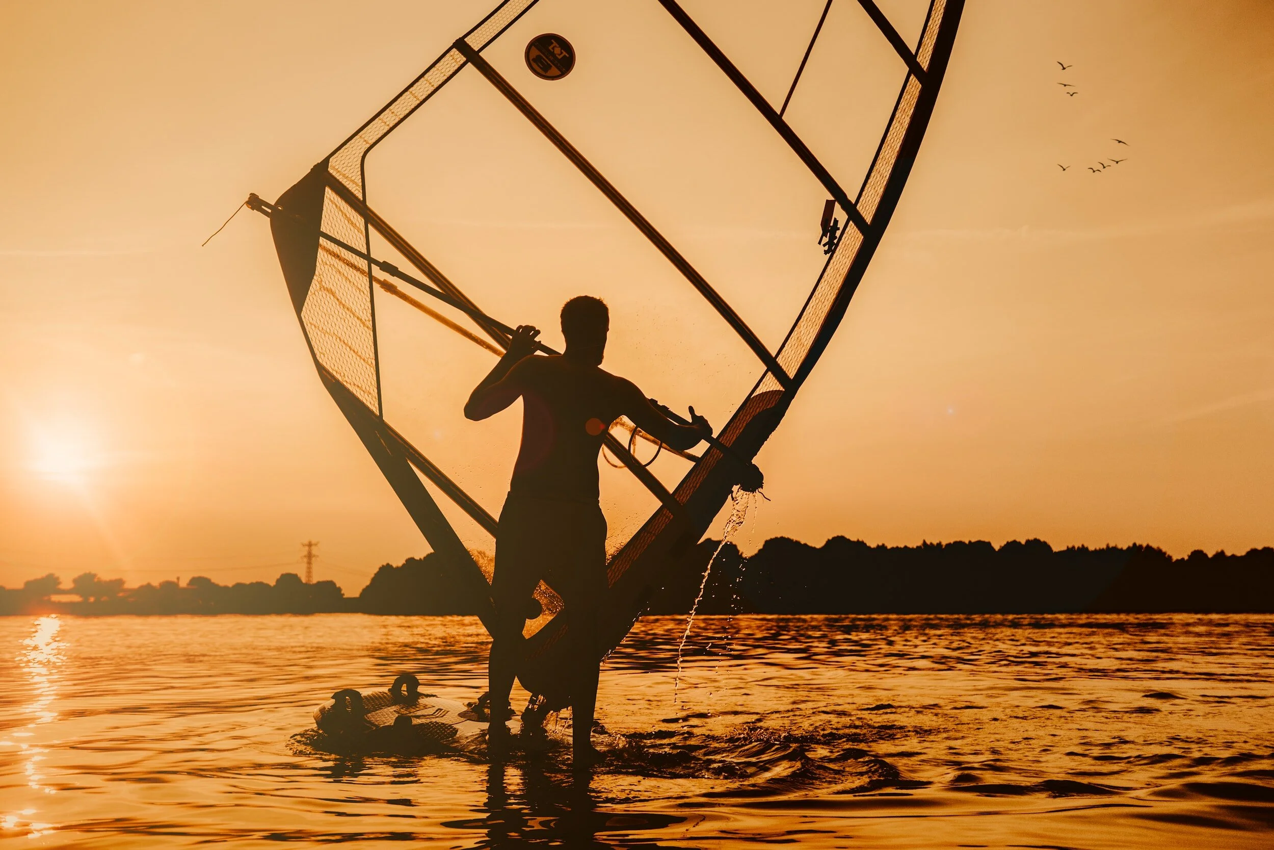 Silhouette of a person holding a windsurfing sail against a setting sun over a body of water.