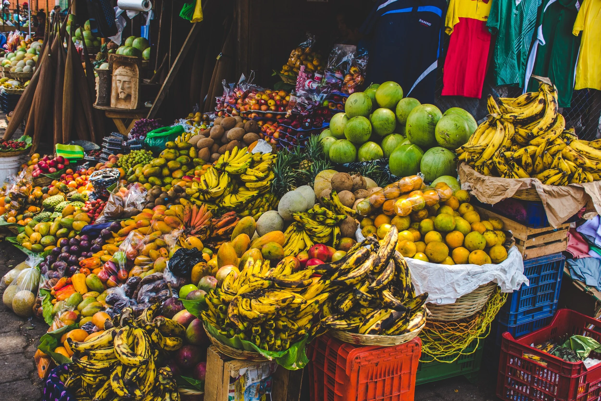 Fresh fruits at an outdoor market, including bananas, watermelons, apples, kiwis, pineapples, and other tropical fruits displayed in baskets and on tables.