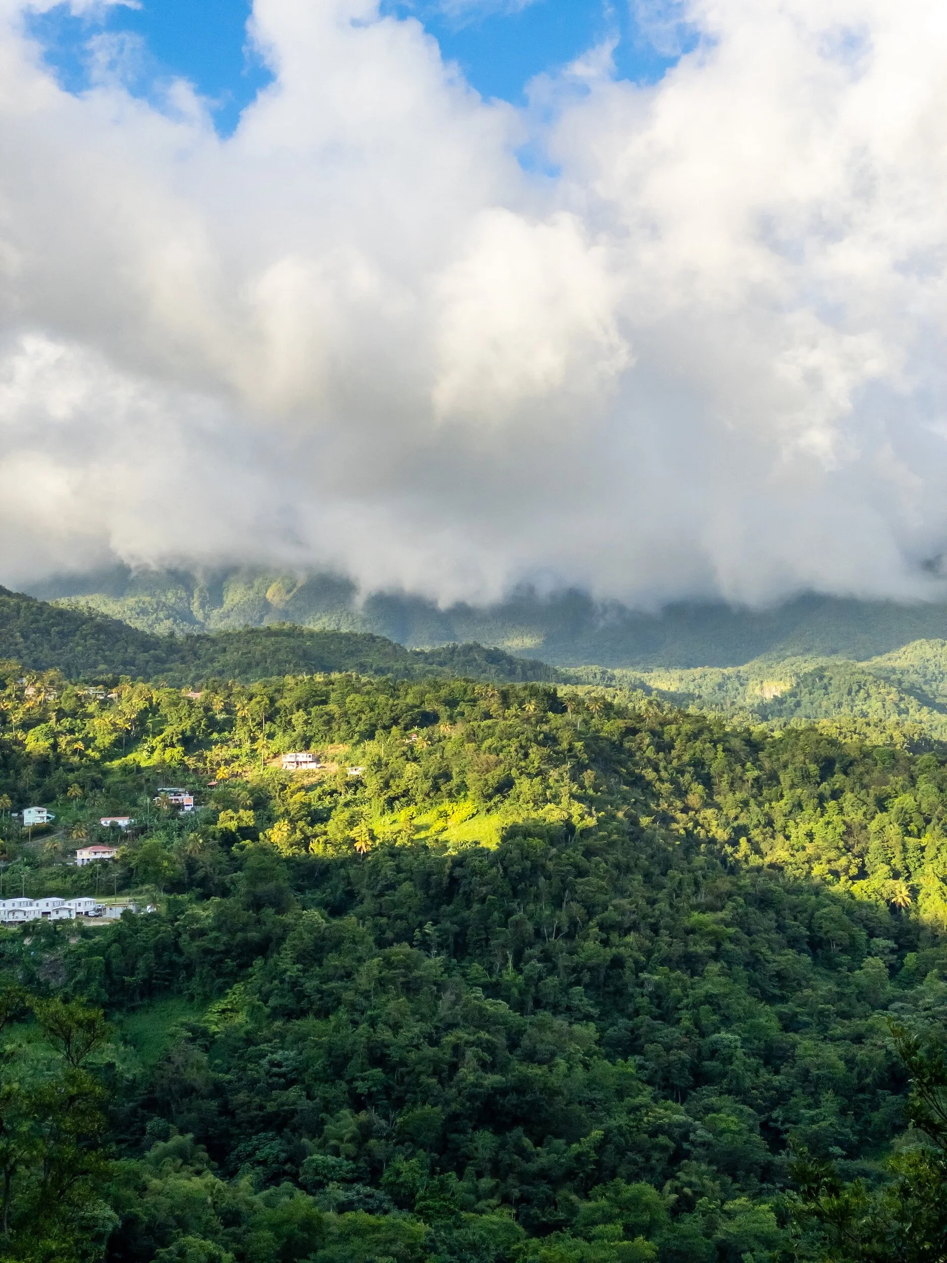 Lush green hillside with scattered houses, clouds over mountains in the background, and a partly cloudy sky.