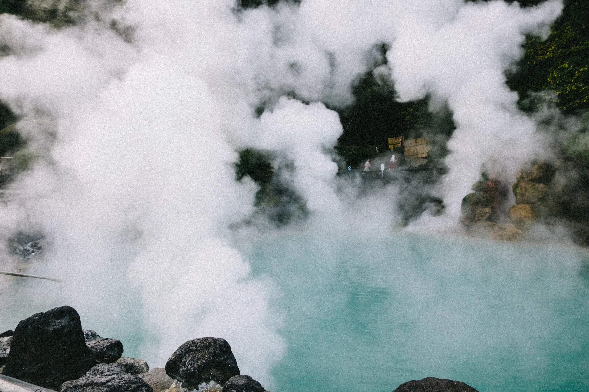 Hot spring with steam rising over turquoise water, surrounded by rocks and greenery.