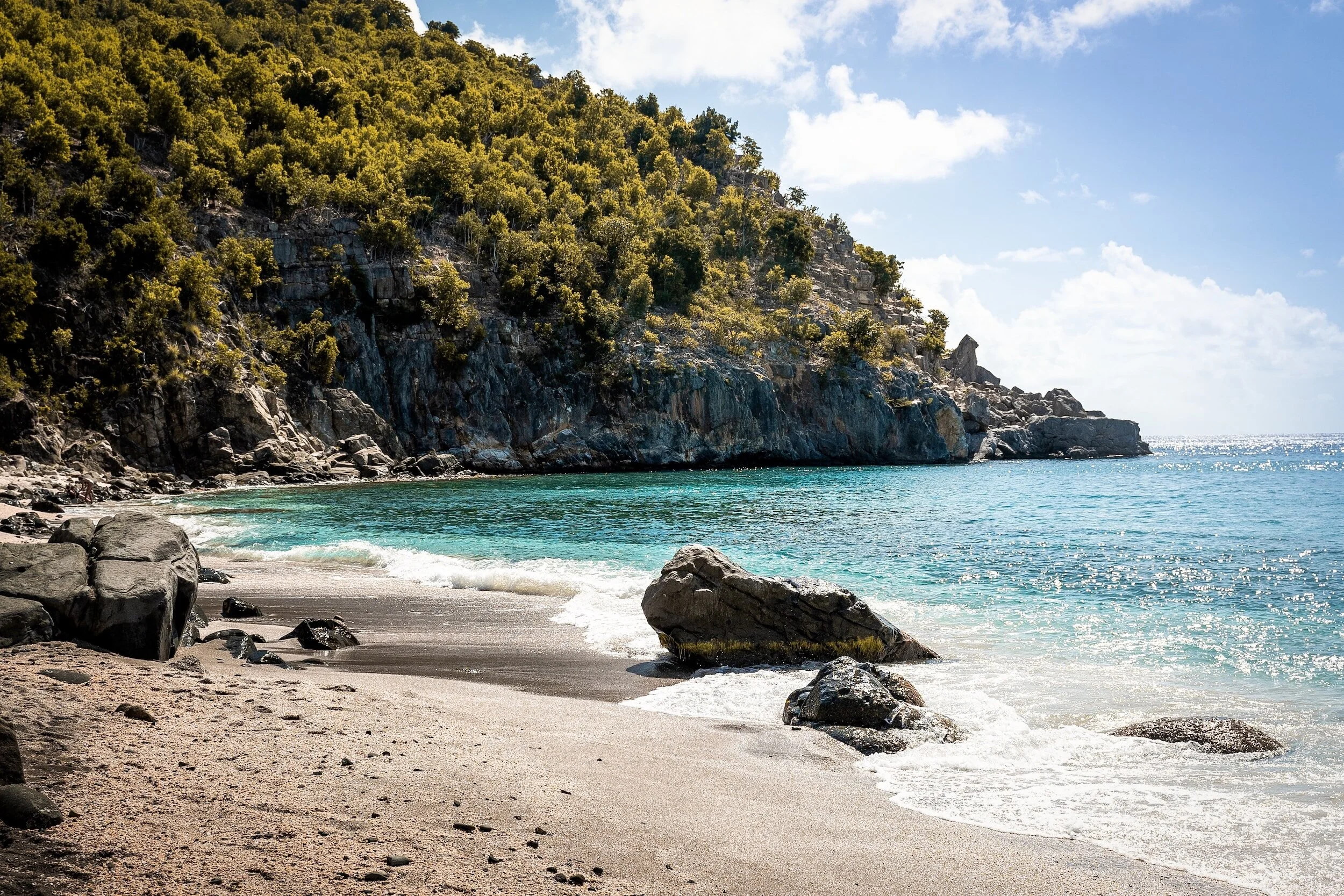 A scenic beach with sandy shore, turquoise water, rocky cliffs covered in green vegetation, and a partly cloudy sky.