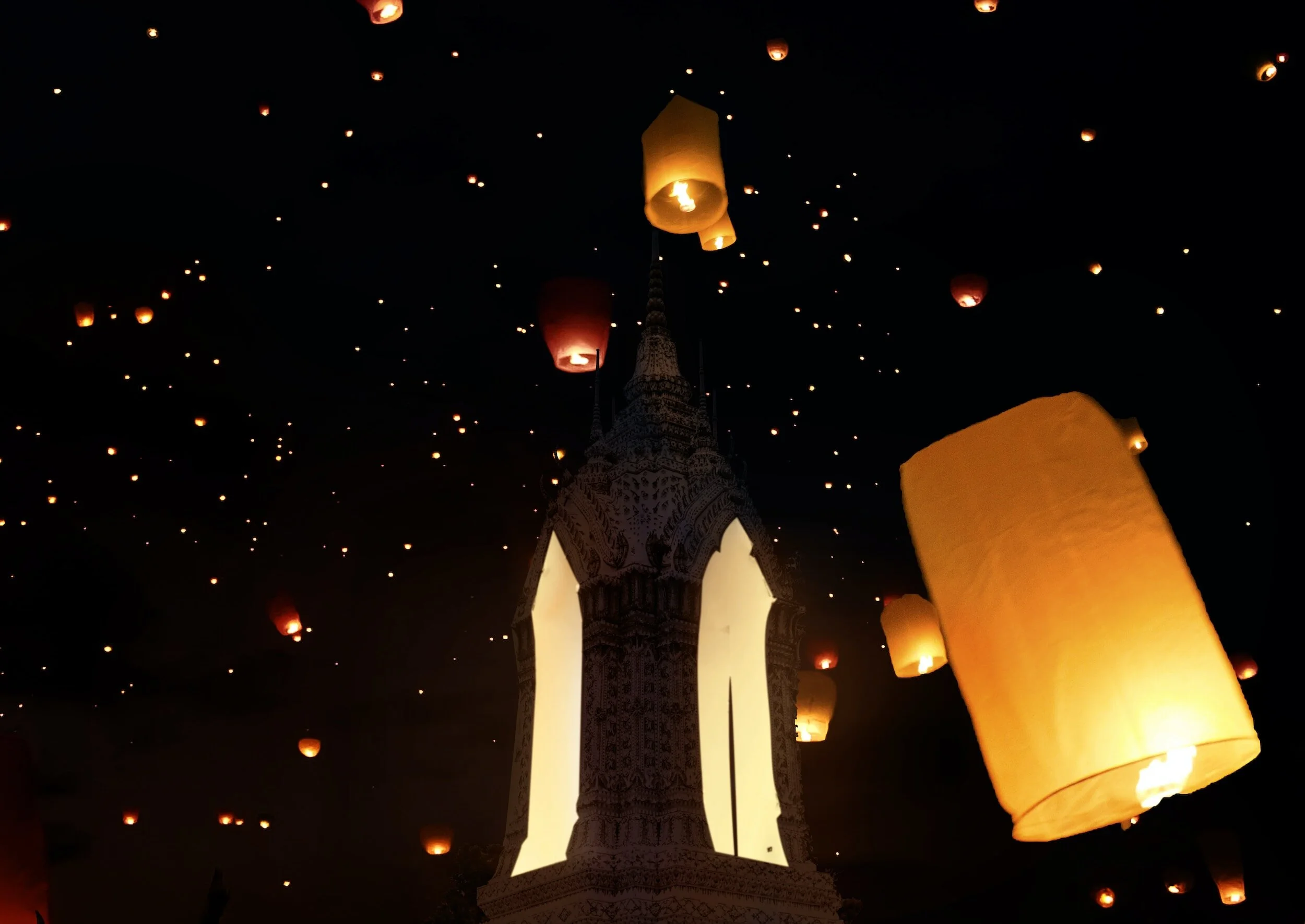 Numerous floating sky lanterns glowing orange and yellow in the dark night sky, with a temple or shrine structure visible in the foreground.