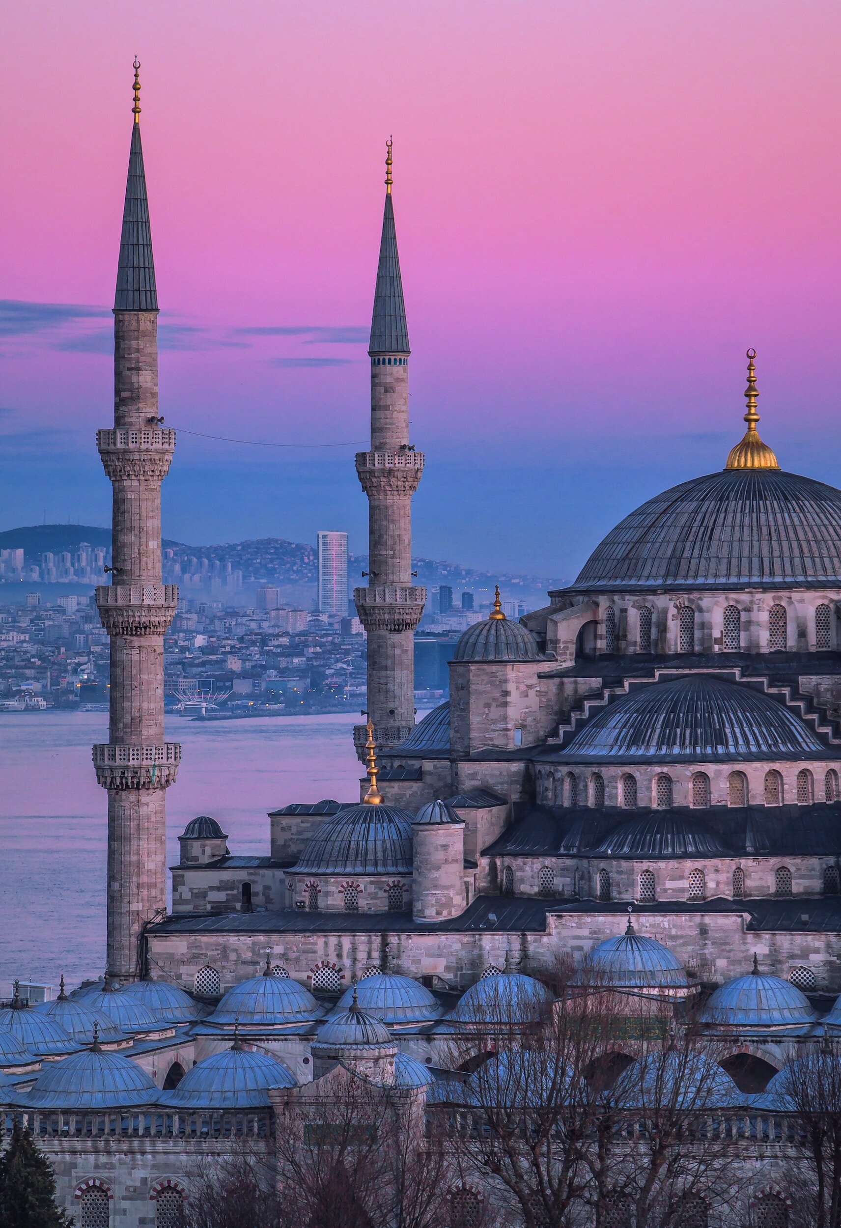 A mosque with two tall minarets and multiple domes under a colorful pink and purple sky, with a cityscape and water in the background.