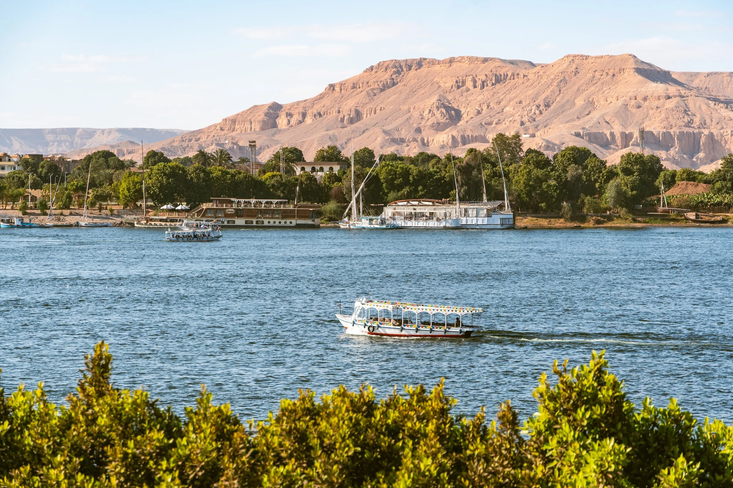 Boats sailing on the Nile River with green trees along the bank and arid mountains in the background.