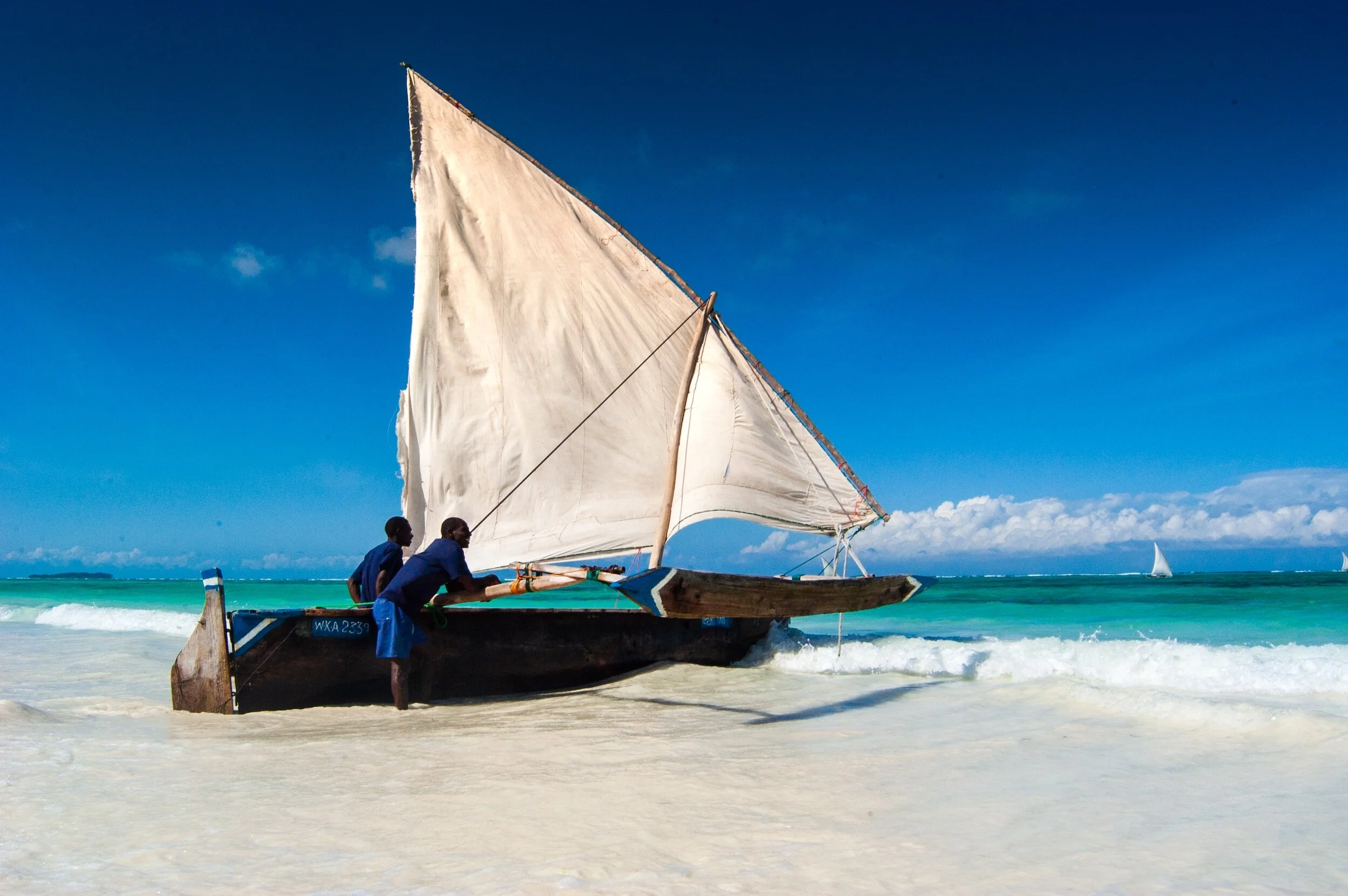 Two men push a traditional sailboat onto the sandy beach as the ocean waves roll in, with a bright blue sky overhead and a smaller sailboat visible on the water in the background.