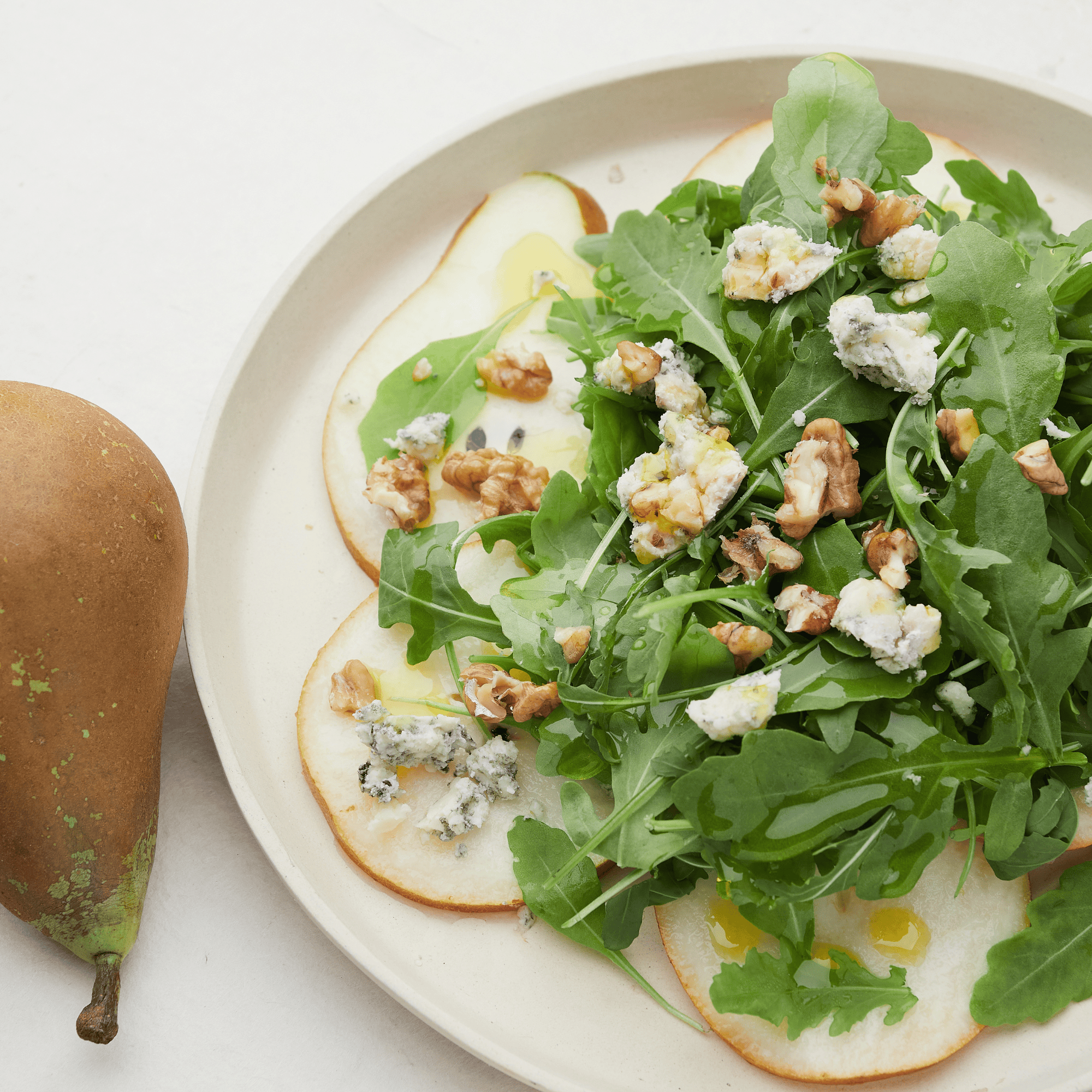 Carpaccio de poire avec roquefort émietté, roquette fraîche et cerneaux de noix, servi dans une assiette blanche