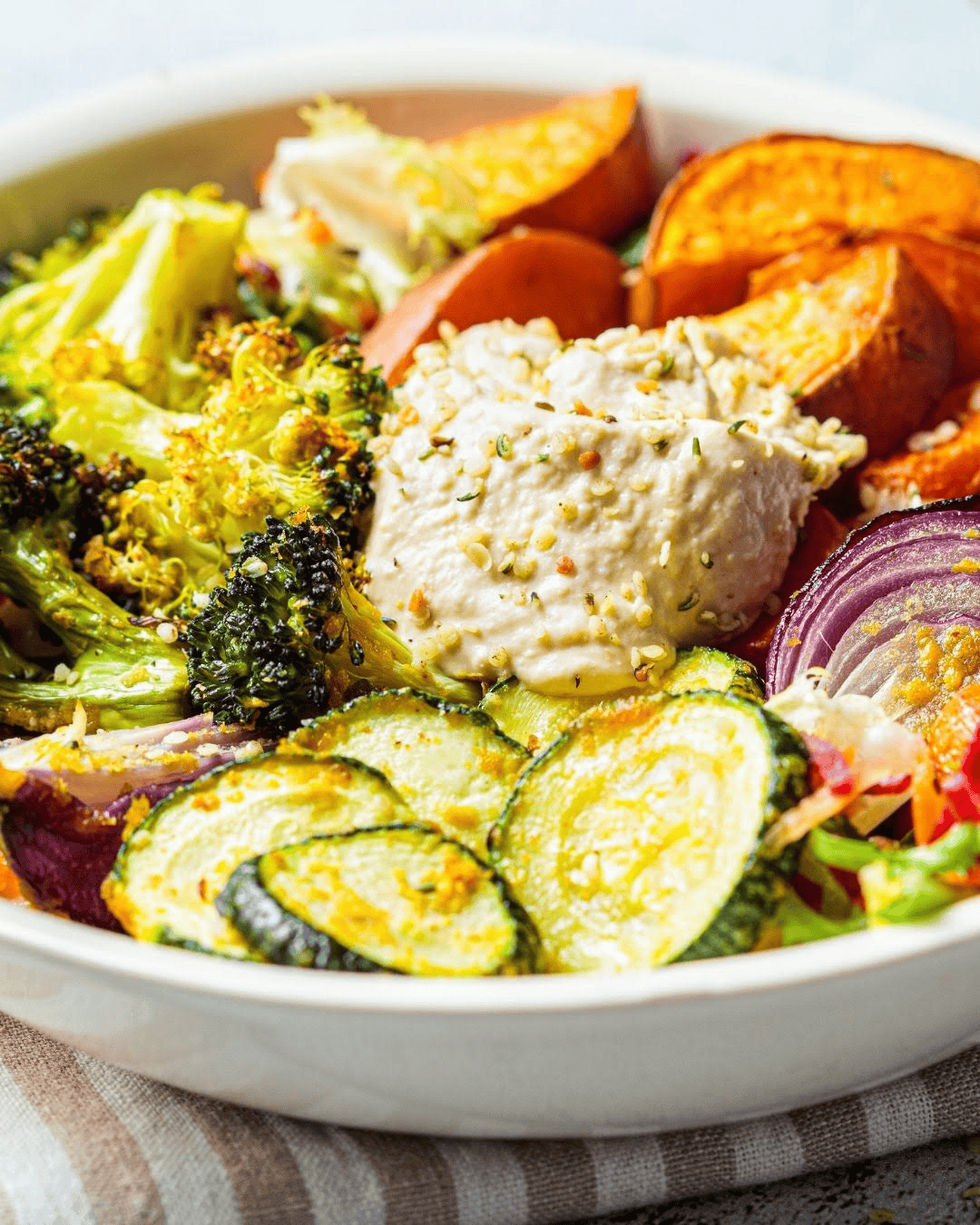 Bol coloré avec légumes rôtis au four (patate douce, brocoli, carottes, oignon rouge) et houmous crémeux maison au centre, parsemé de graines
