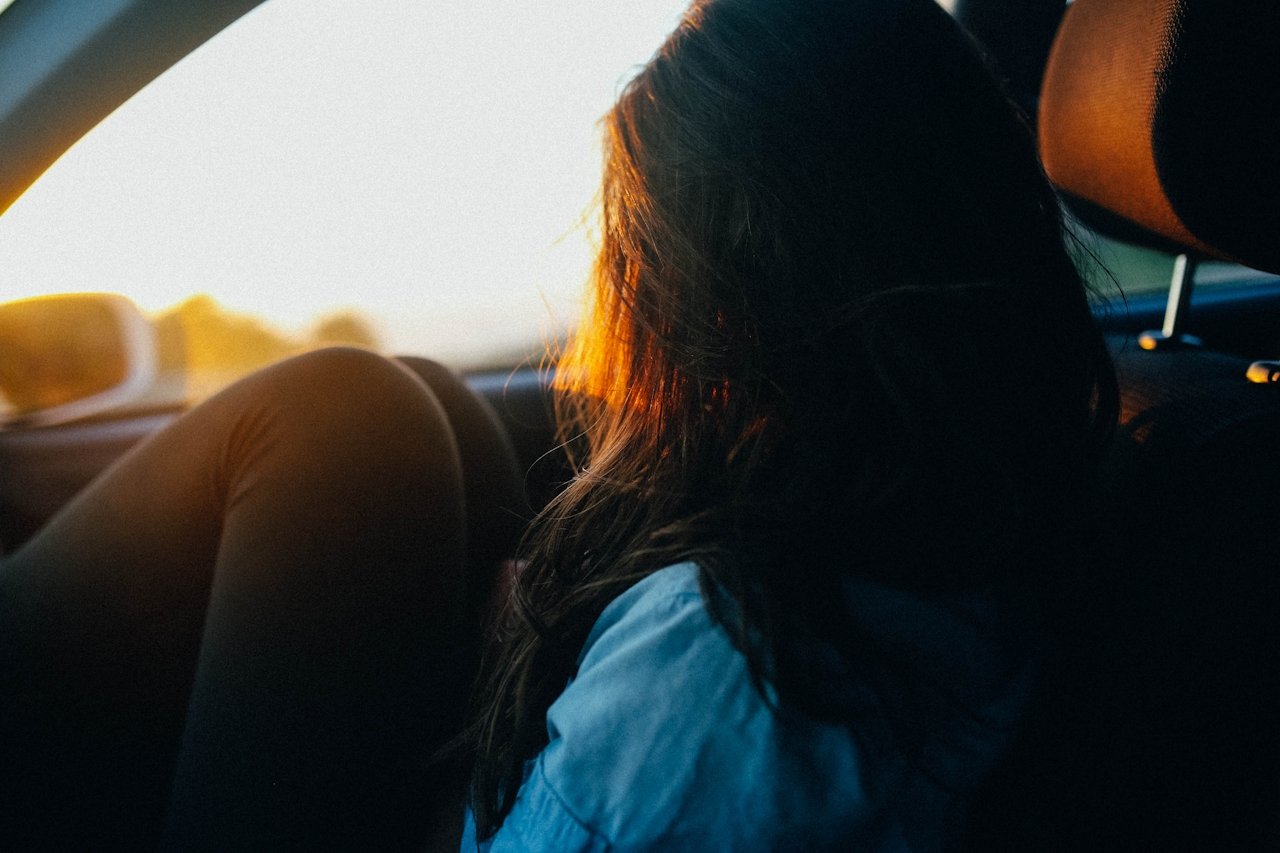 A woman with dark hair resting inside a car during sunset, looking out the window.