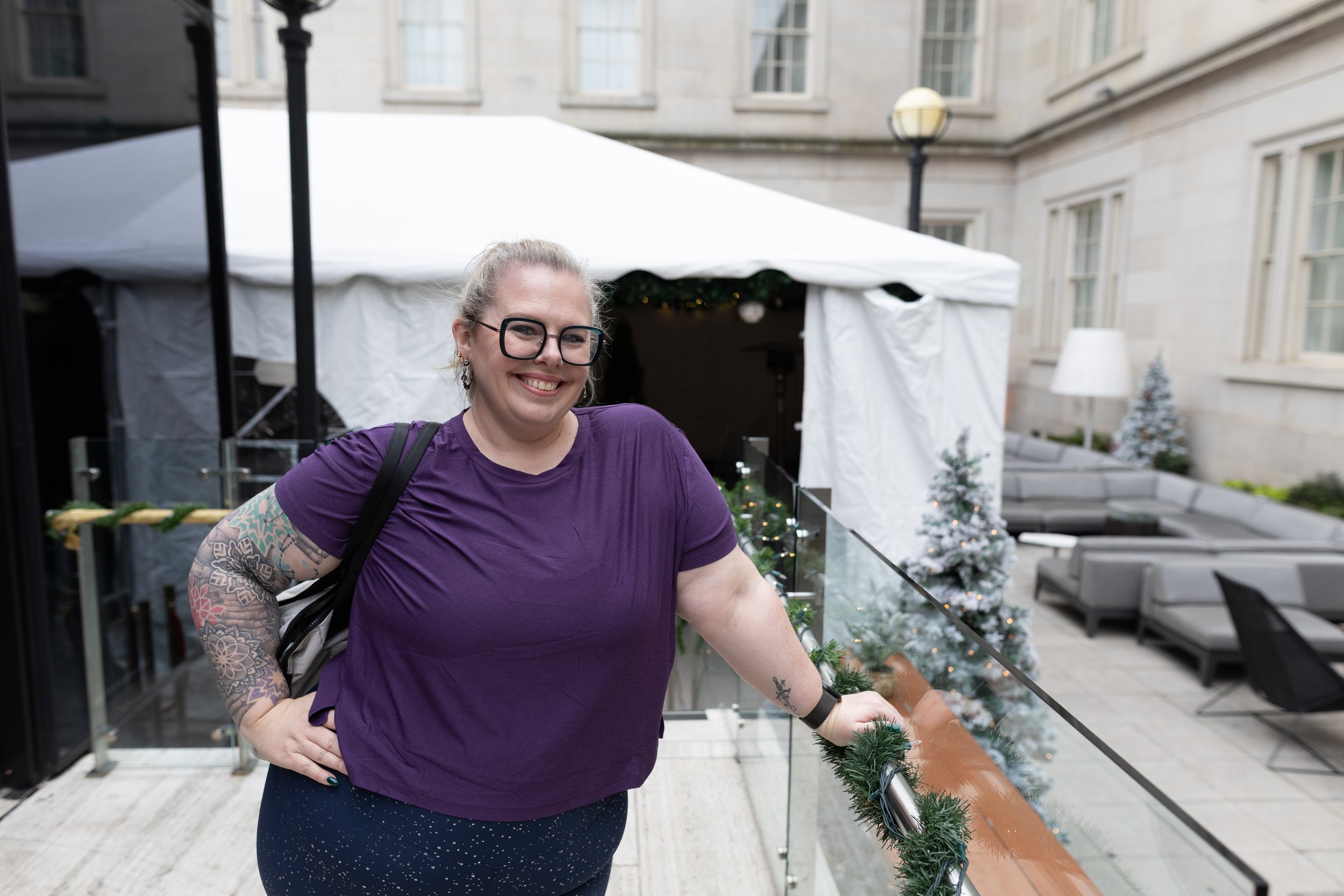 A smiling woman with glasses, wearing a purple shirt and dark blue pants, standing on an outdoor balcony decorated with Christmas garland and small Christmas trees, in front of a white tent outside a building.