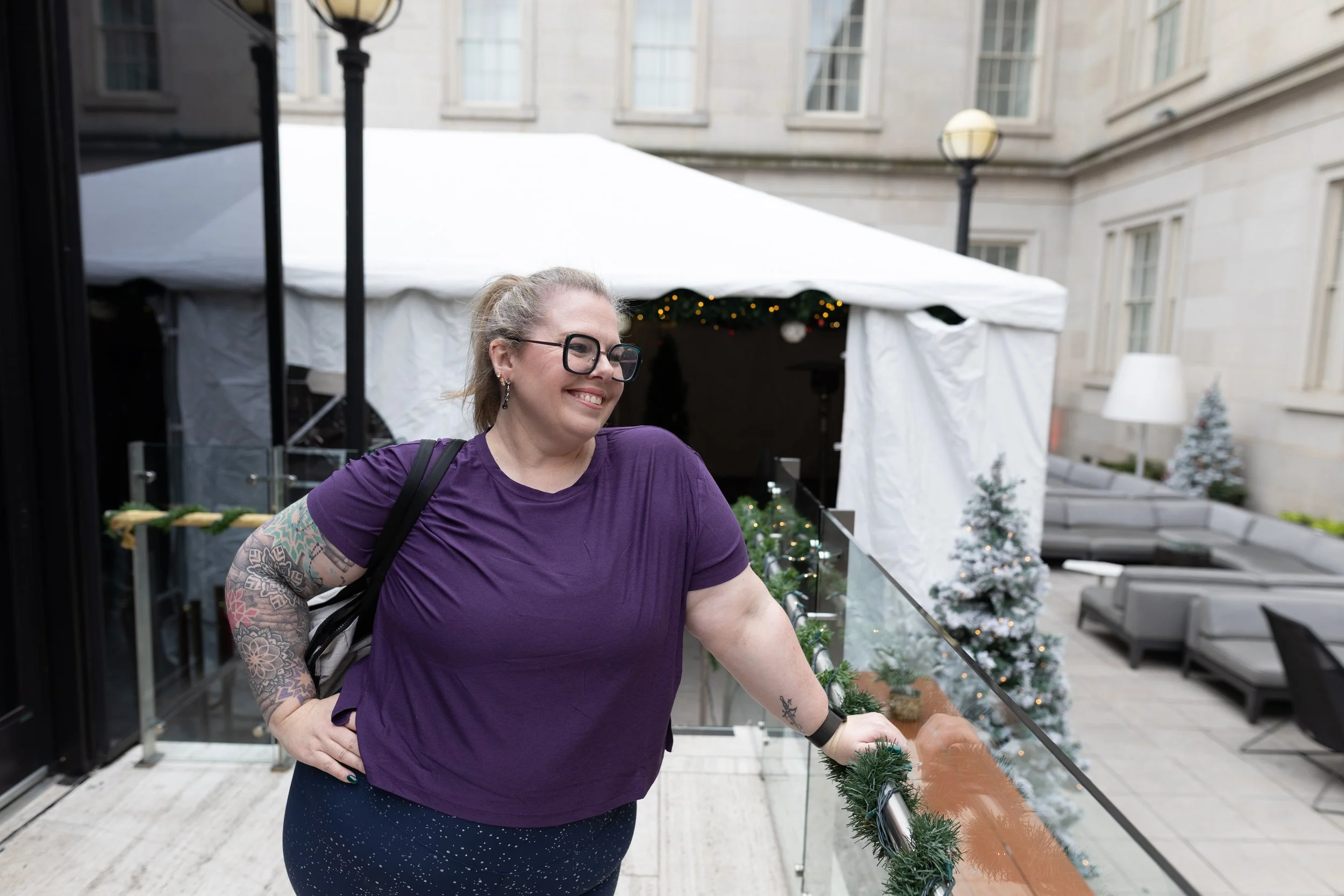 A woman with glasses, tattoos on her arm, and a purple shirt, smiling and leaning on a decorated railing on a rooftop patio with Christmas trees and a tent in the background.