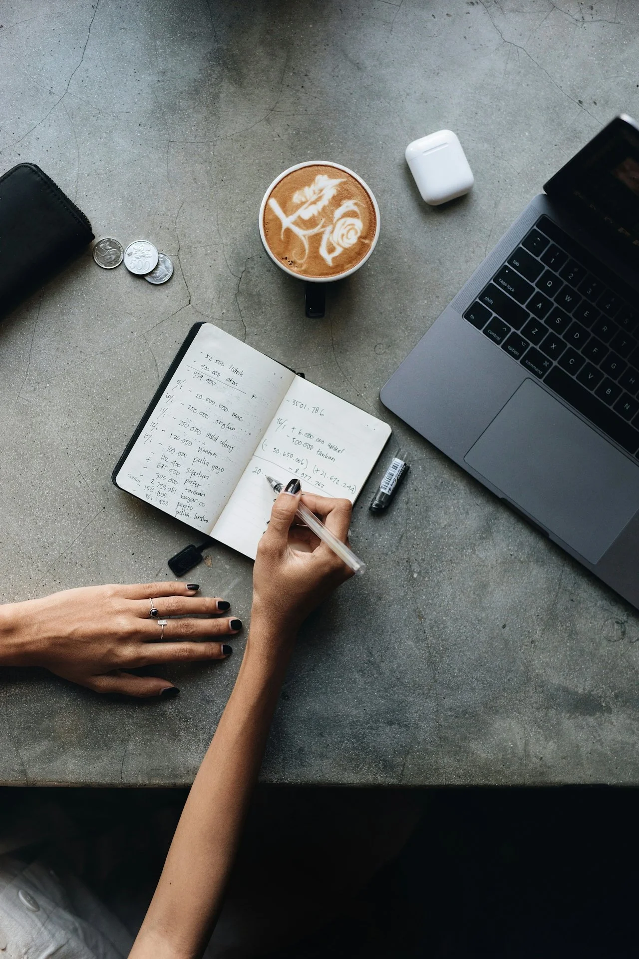 A person writing notes in a notebook on a table with a laptop, a cup of coffee with latte art, a pack of wireless earphones, some coins, a tablet, and a black wallet.