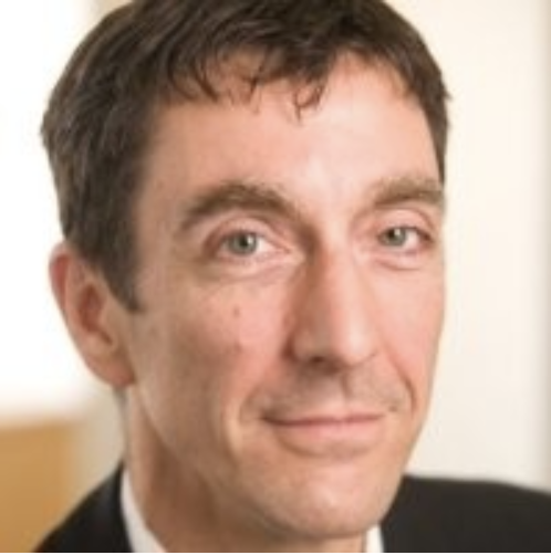Close-up photo of a man with short dark hair and light skin, wearing a suit and tie, smiling slightly.