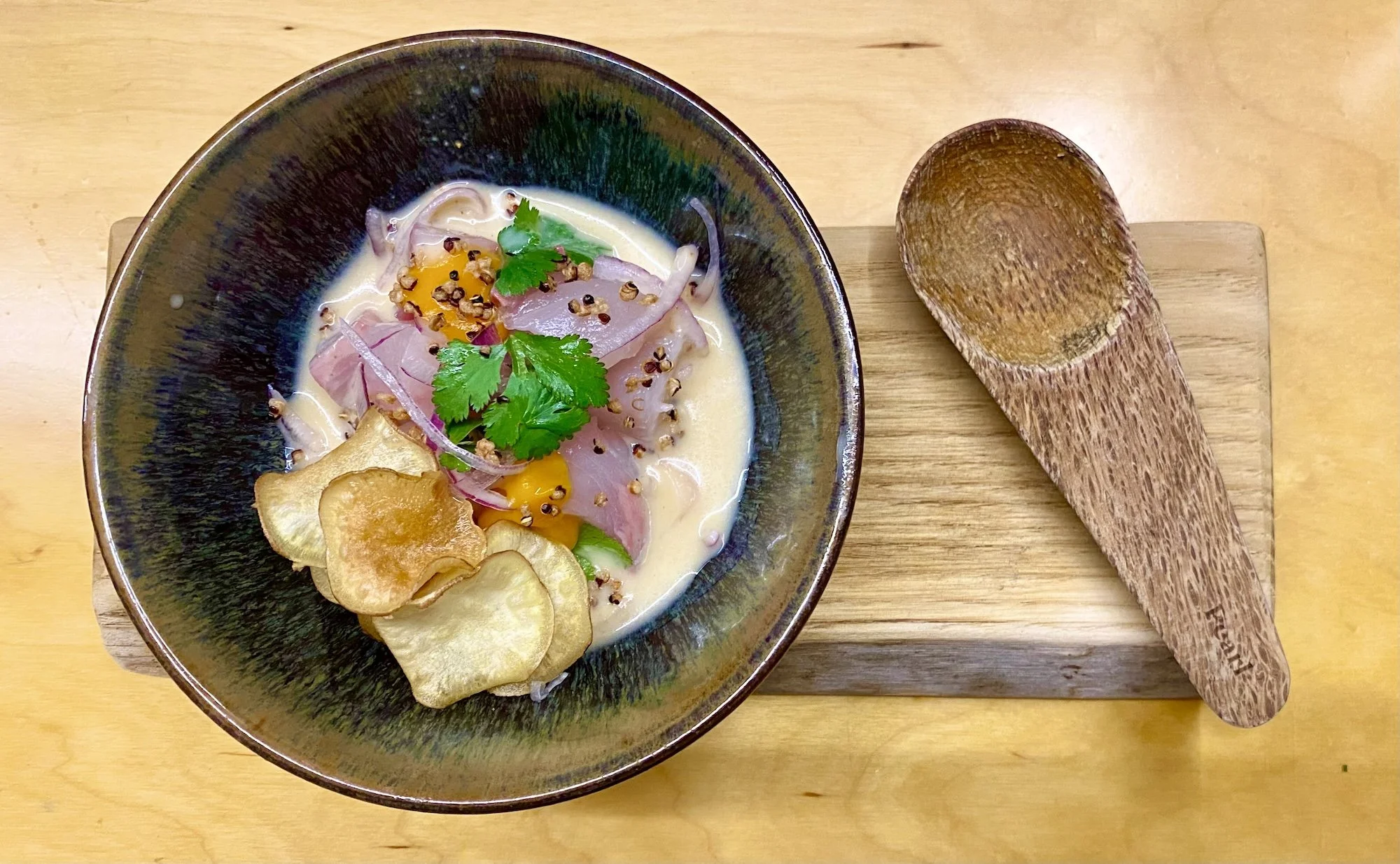 A bowl of ceviche garnished with cilantro, red onion, and sliced plantain chips, on a wooden board with a wooden spoon.