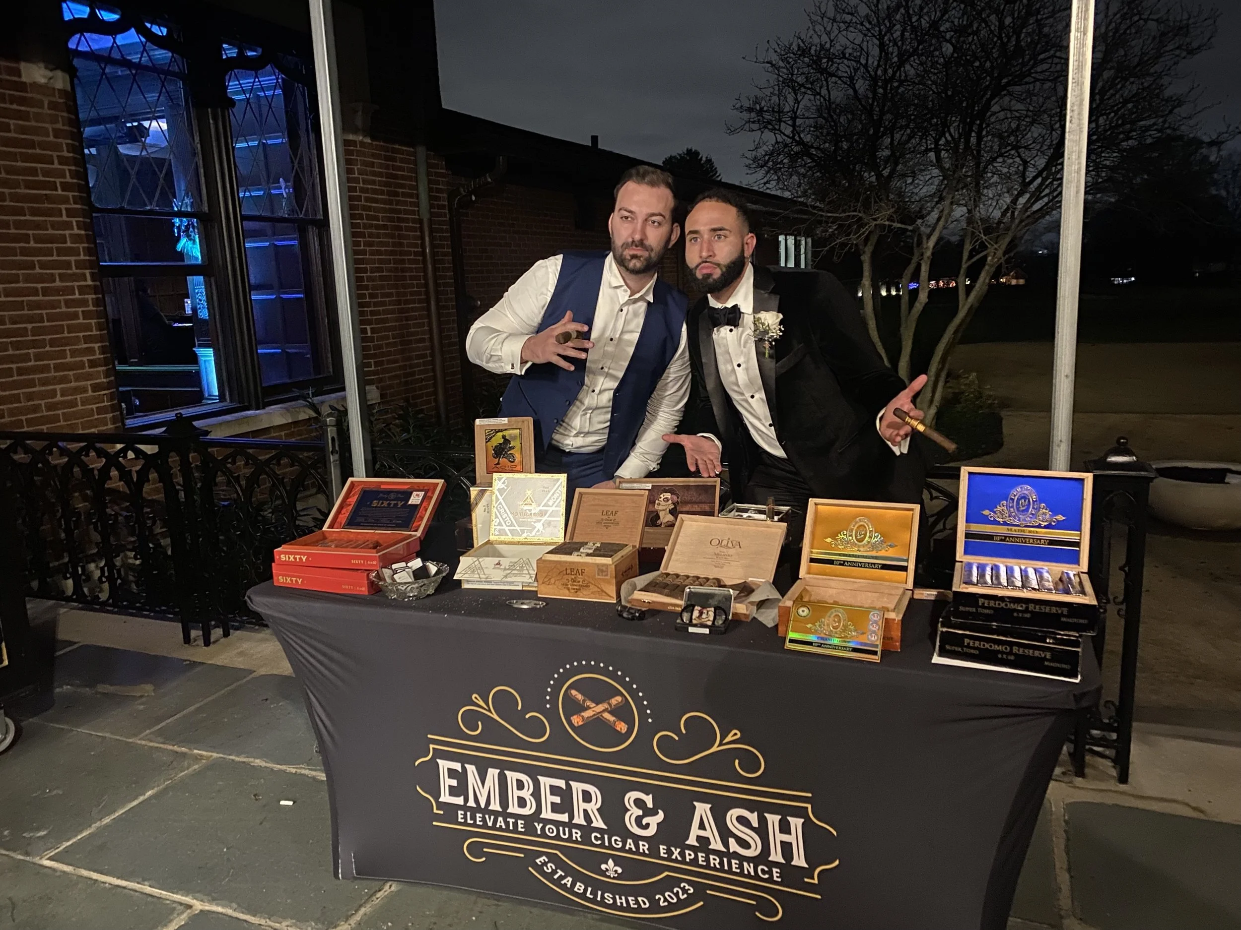 Two men in formal attire standing behind a table at a cigar event, with boxes of cigars on display, outdoors at night.
