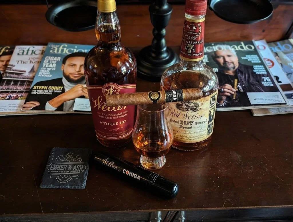 Two bottles of bourbon, a glass of bourbon, a cigar resting on the bottles, magazines on a wooden table, a cigar lighter, and a coaster for an ashtray.