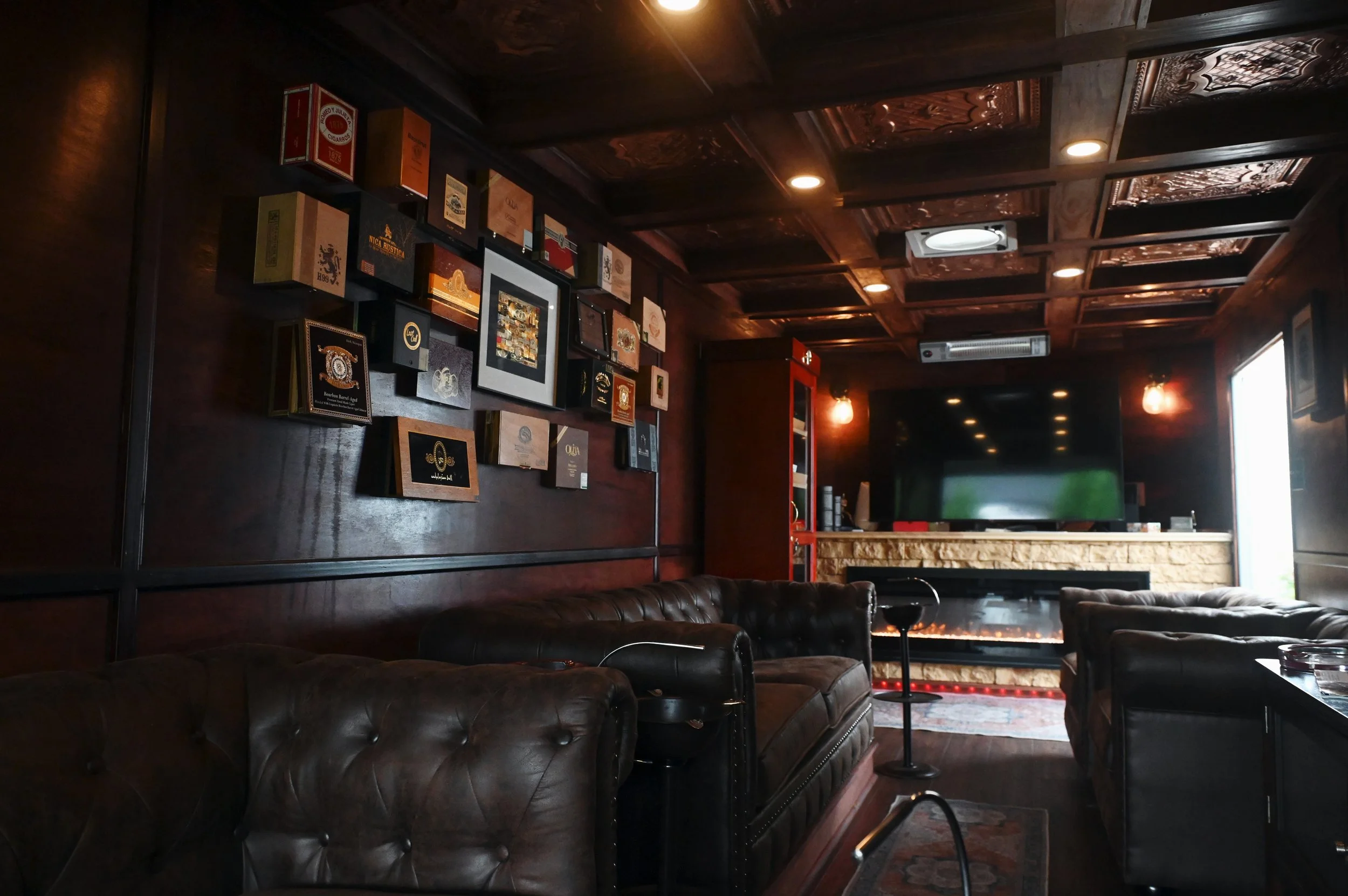 Cozy lounge with dark wooden paneling, leather sofas, wall art, and a large flat-screen TV above a fireplace.