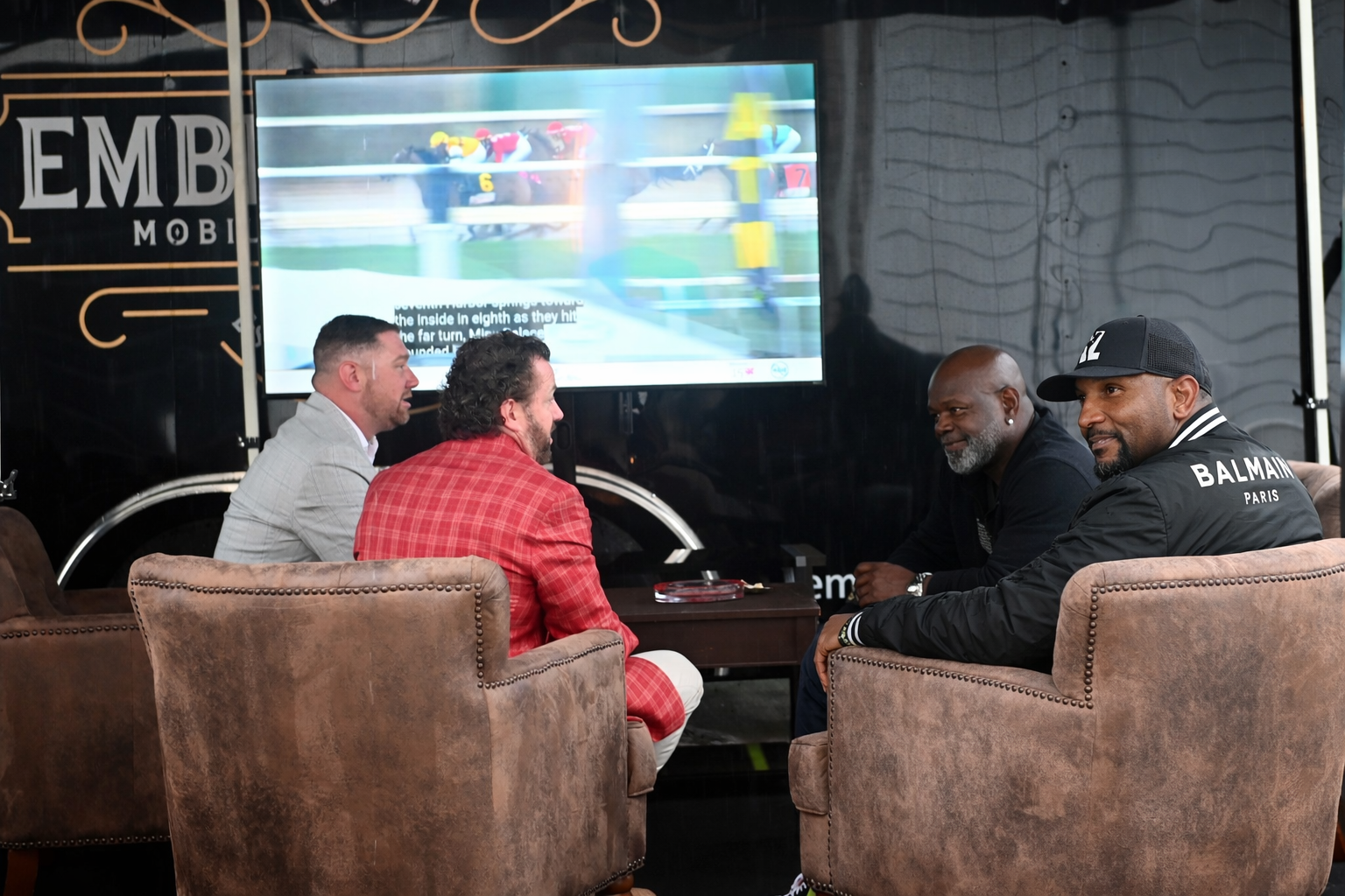 Four men sit on brown armchairs watching a horse race on a large screen in a dark room. The screen shows horses racing on a track.