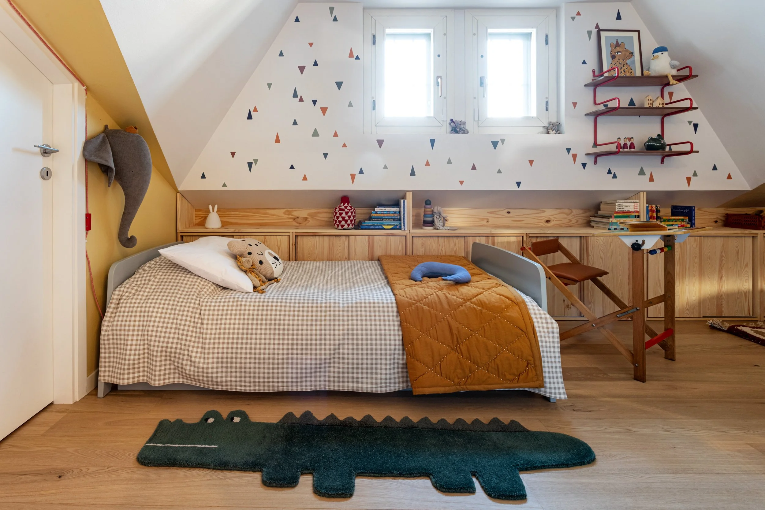 Camera mansardata per bambini con letto, parete decorata e scrivania sotto le finestre.