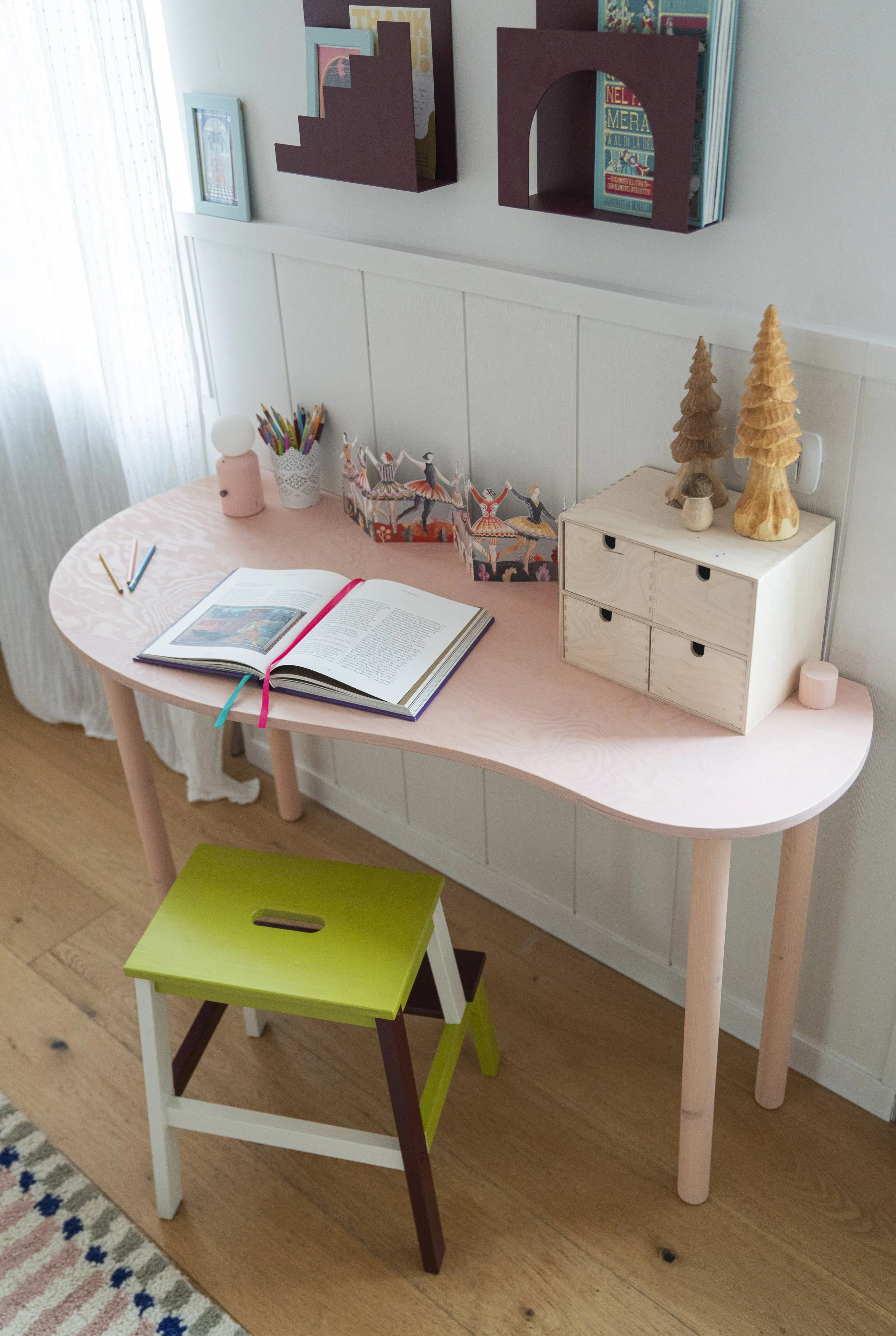 Scrivania rosa per bambini con sedia verde, libri e contenitori per disegno e creatività.