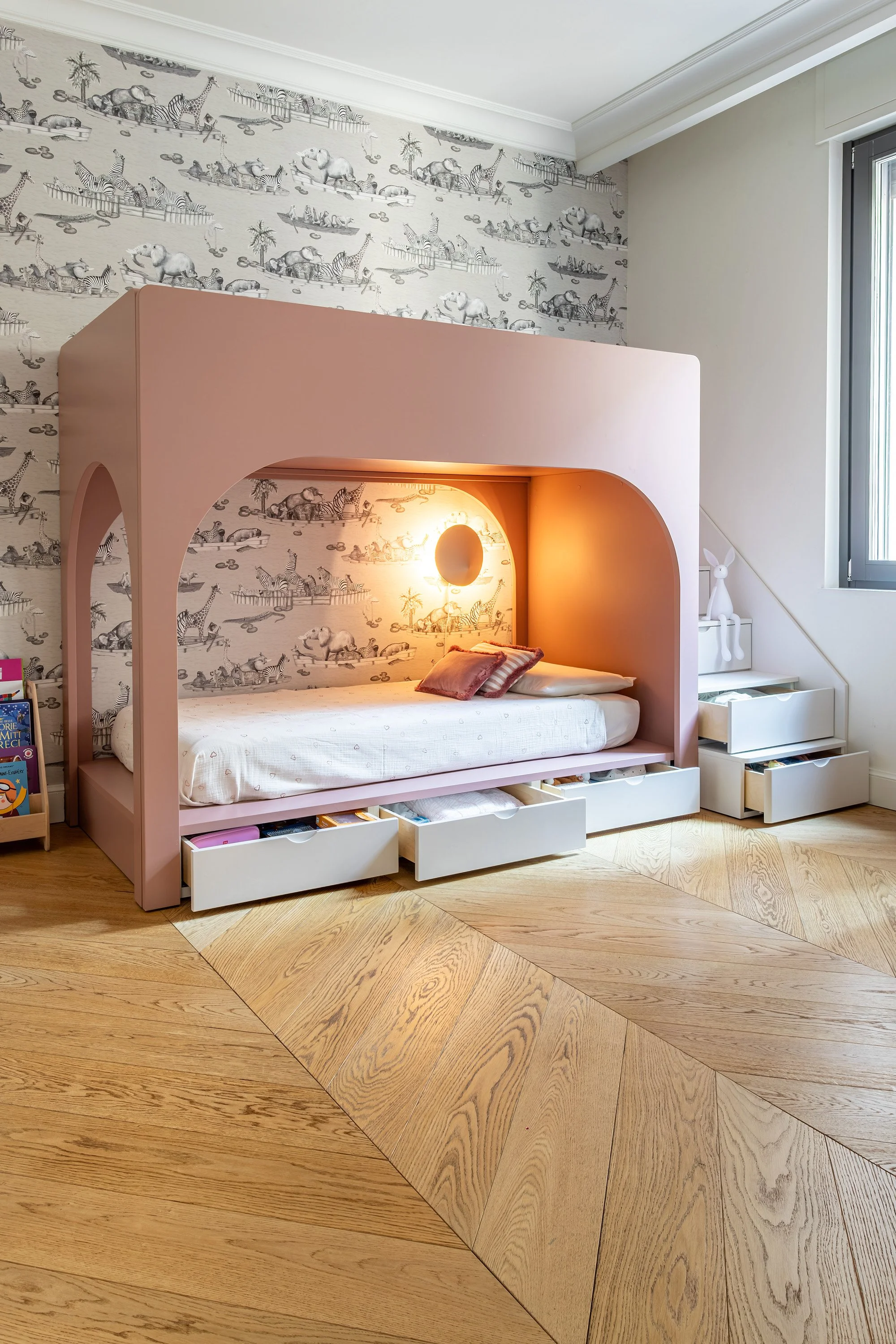 Letto inserito in una nicchia rosa ad arco, con carta da parati illustrata, contenitori inferiori e scala laterale.