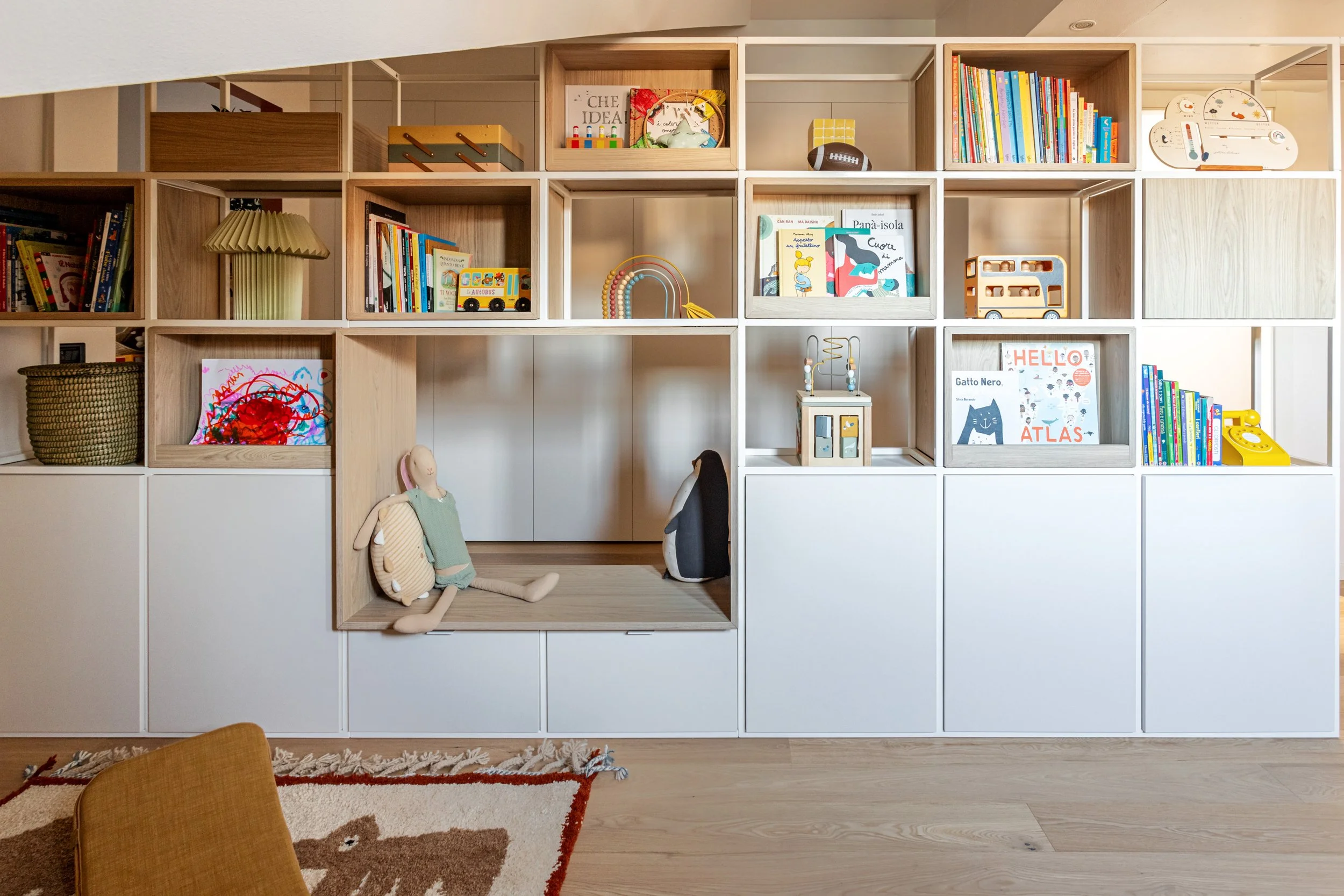 Parete-libreria passante con panca integrata e vani contenitore accessibili da entrambi i lati.