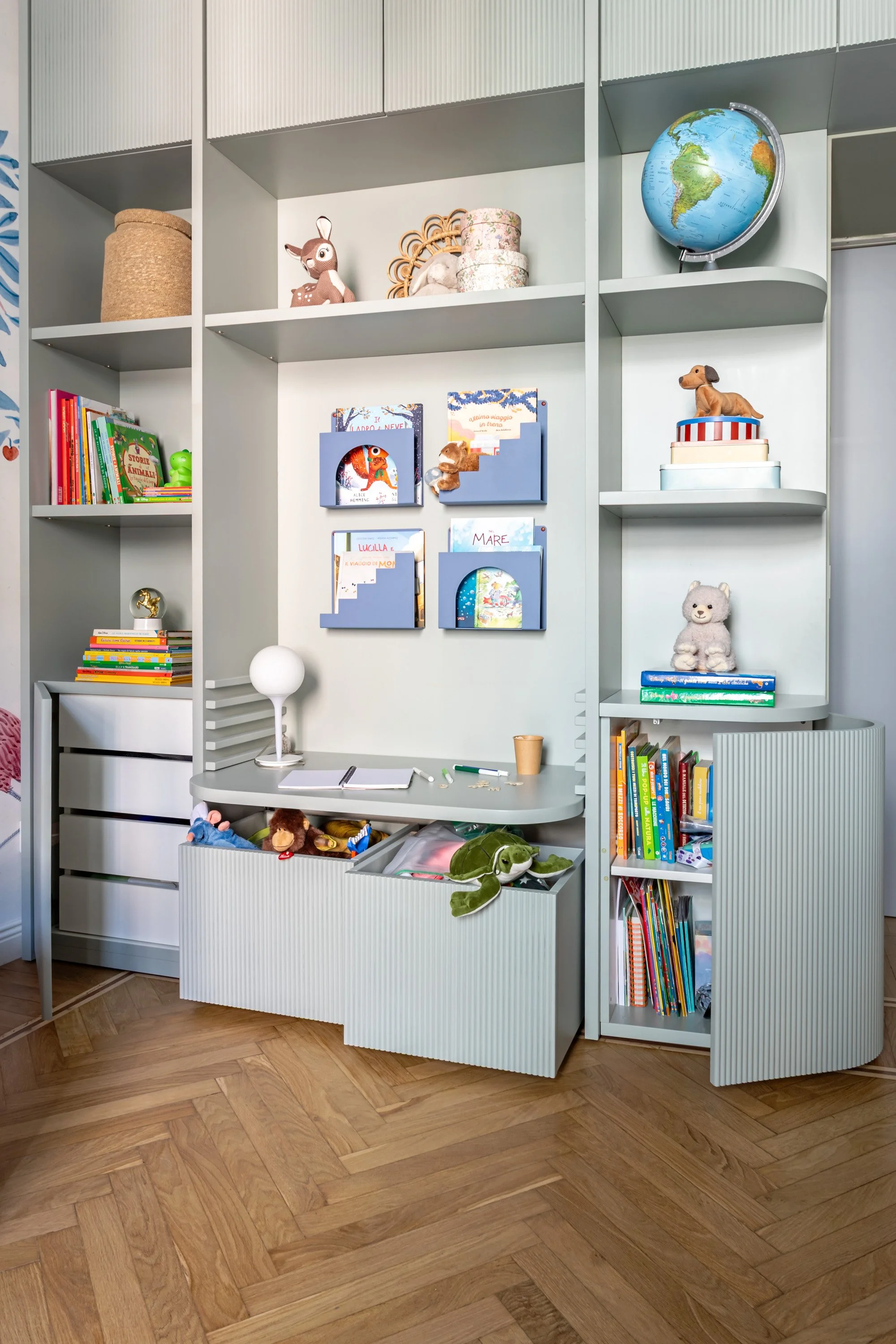 Parete attrezzata con scrivania centrale, librerie laterali e contenitori chiusi per giochi e materiali.