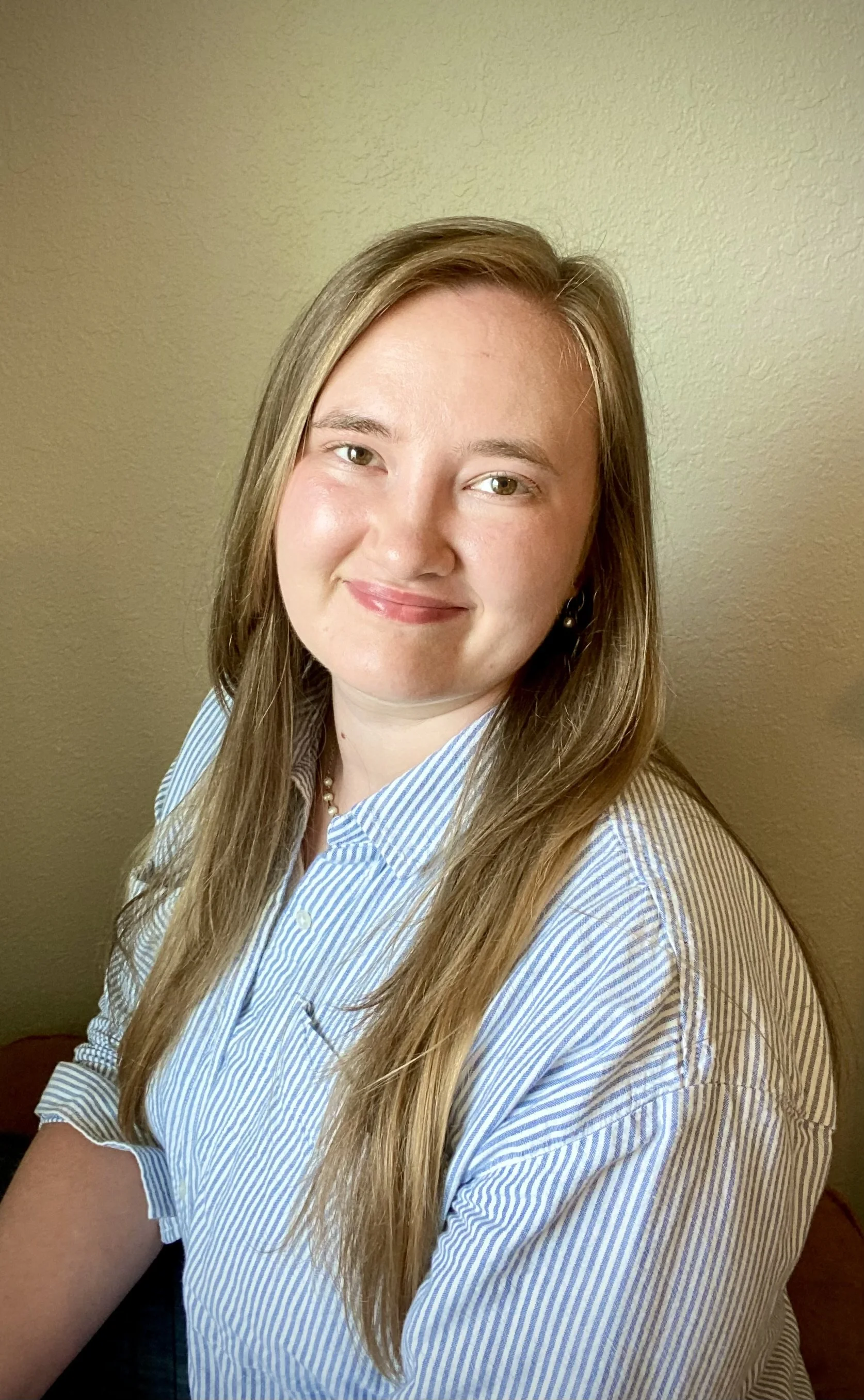 Portrait of a woman with long light brown hair, wearing a blue and white striped button-up shirt, smiling, sitting in a room with beige walls.