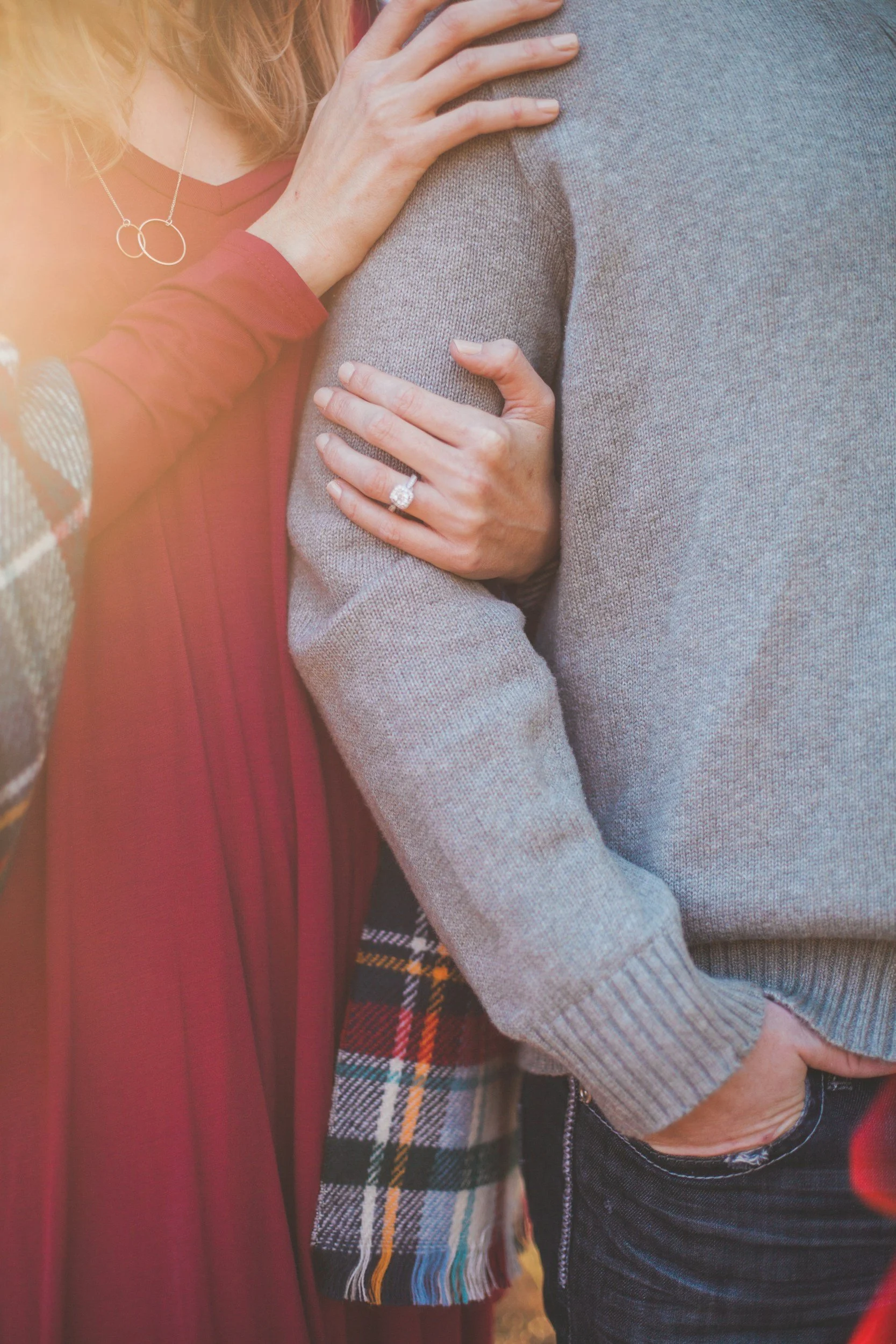 A woman wearing a red dress and a plaid jacket is embracing a man in a gray sweater, with her hand on his chest showing her engagement ring.