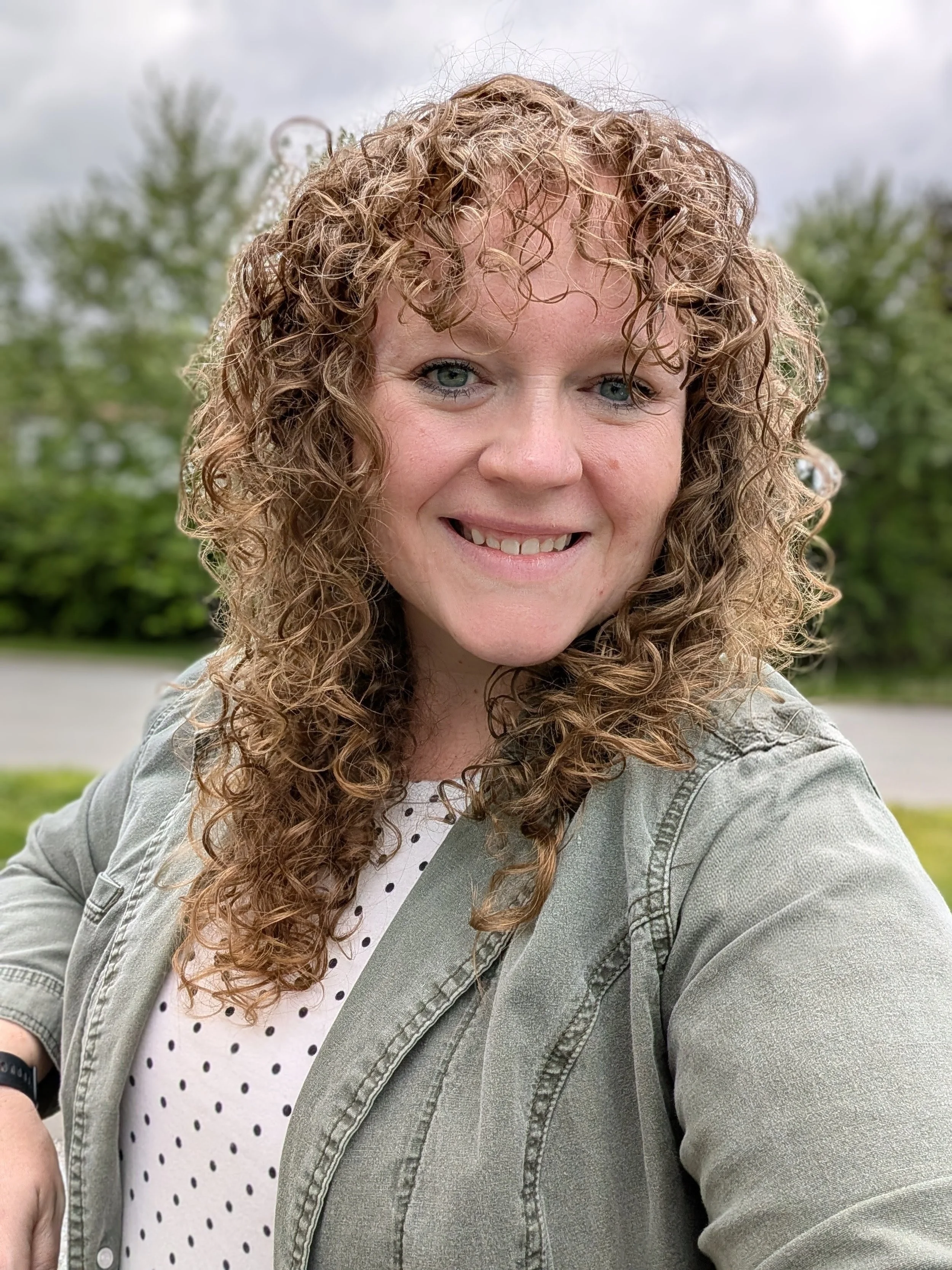 A female marriage counselor with curly red hair smiling outdoors, wearing a polka-dot top and a light jacket, with green trees and a cloudy sky in the background.