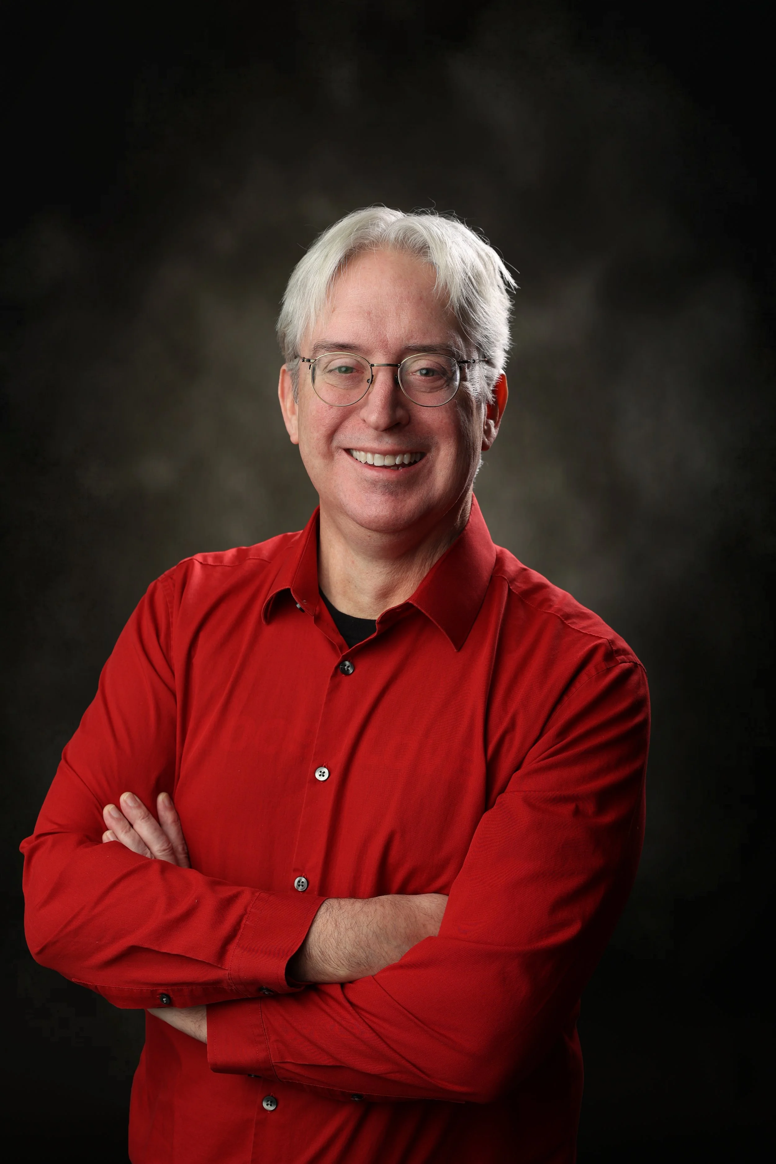 A smiling man with gray hair and glasses, wearing a red shirt, standing with arms crossed against a dark, blurred background.