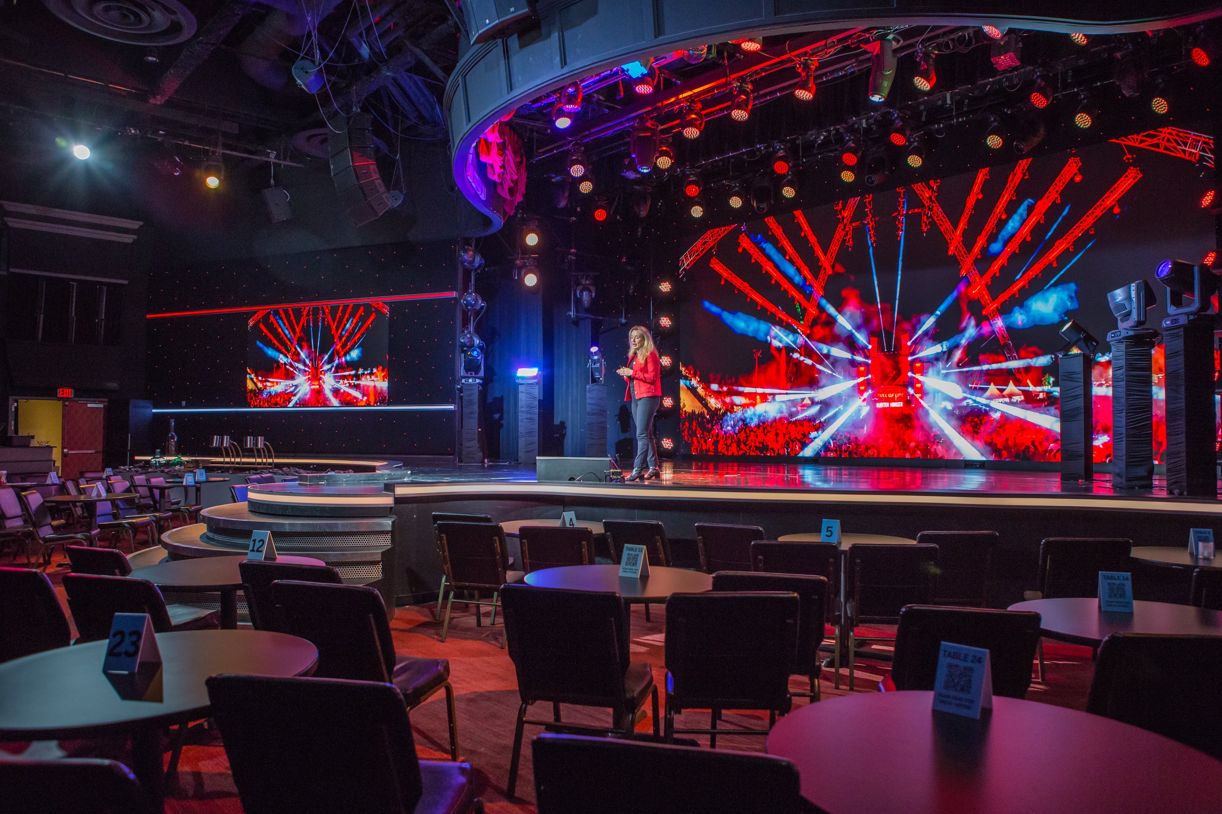 An empty live performance stage with a large LED screen displaying colorful graphics. A woman in a red jacket is standing on stage, speaking into a microphone. Tables and chairs are arranged in front of the stage, with numbered table cards visible. T