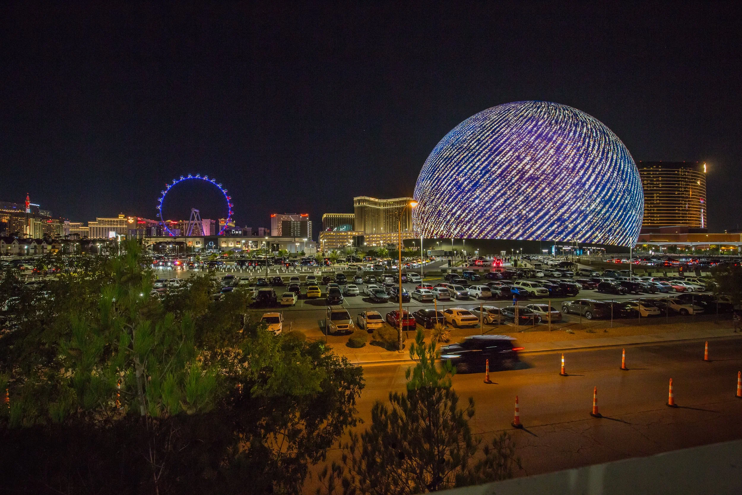 Nighttime view of The Sphere, the Las Vegas skyline, a Ferris wheel, and a parking lot filled with cars.