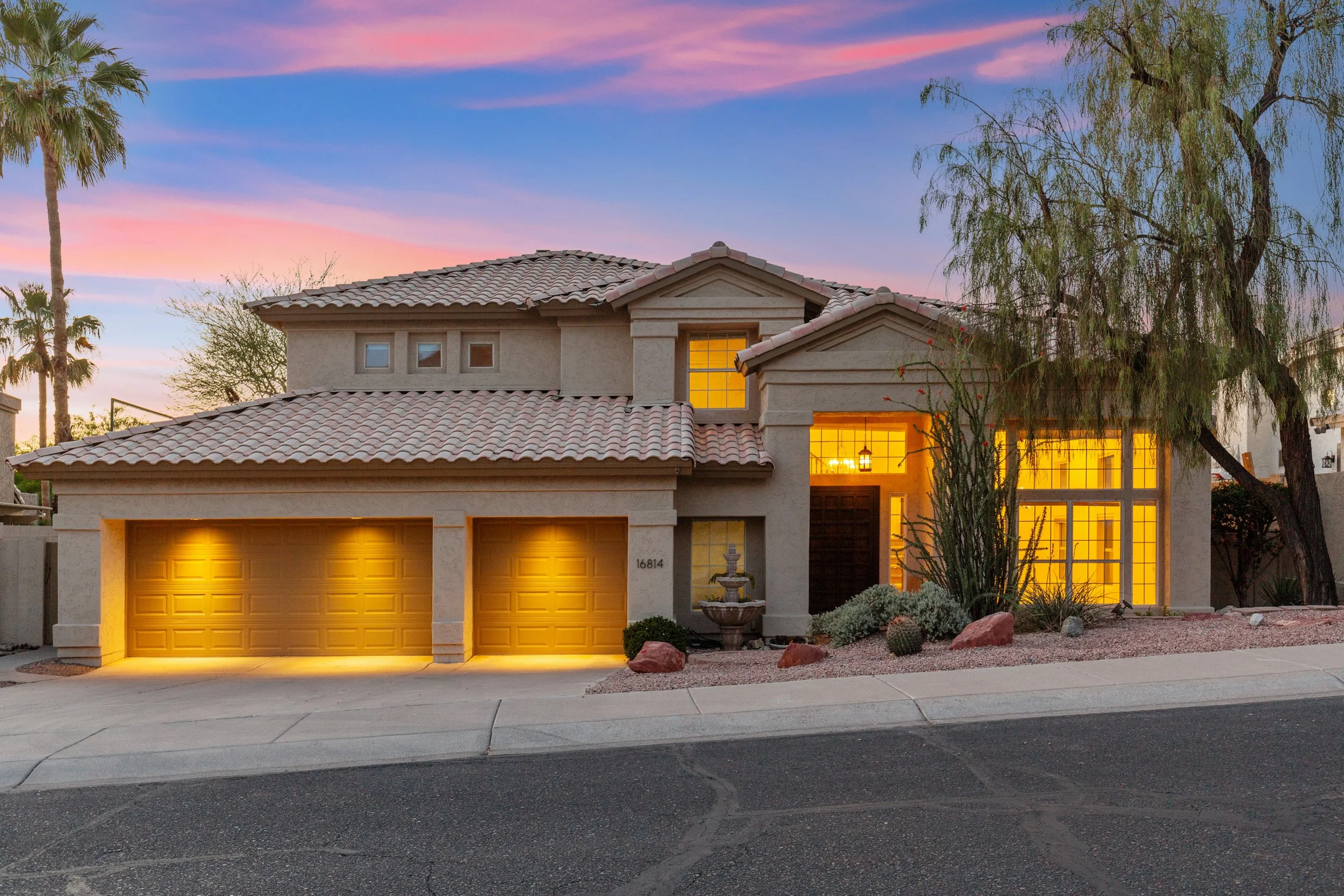 A two-story house with a beige stucco exterior and a tile roof, illuminated from inside at sunset, with a landscaped front yard featuring desert plants and rocks, and a neighborhood street in the foreground.