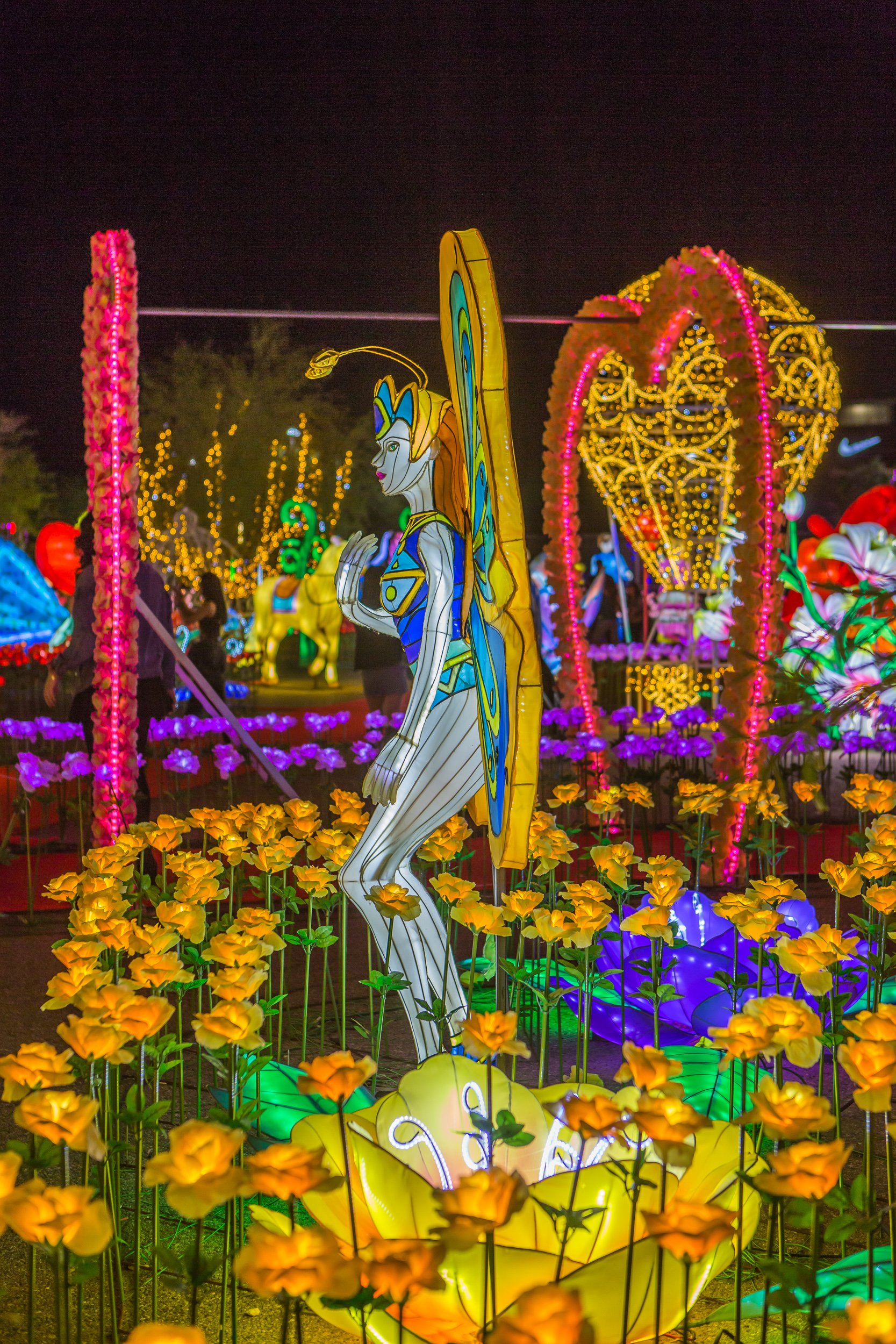 Colorful illuminated lantern display featuring a fairy with butterfly wings, standing amid yellow flower lanterns, with other illuminated figures and decorations in the background.