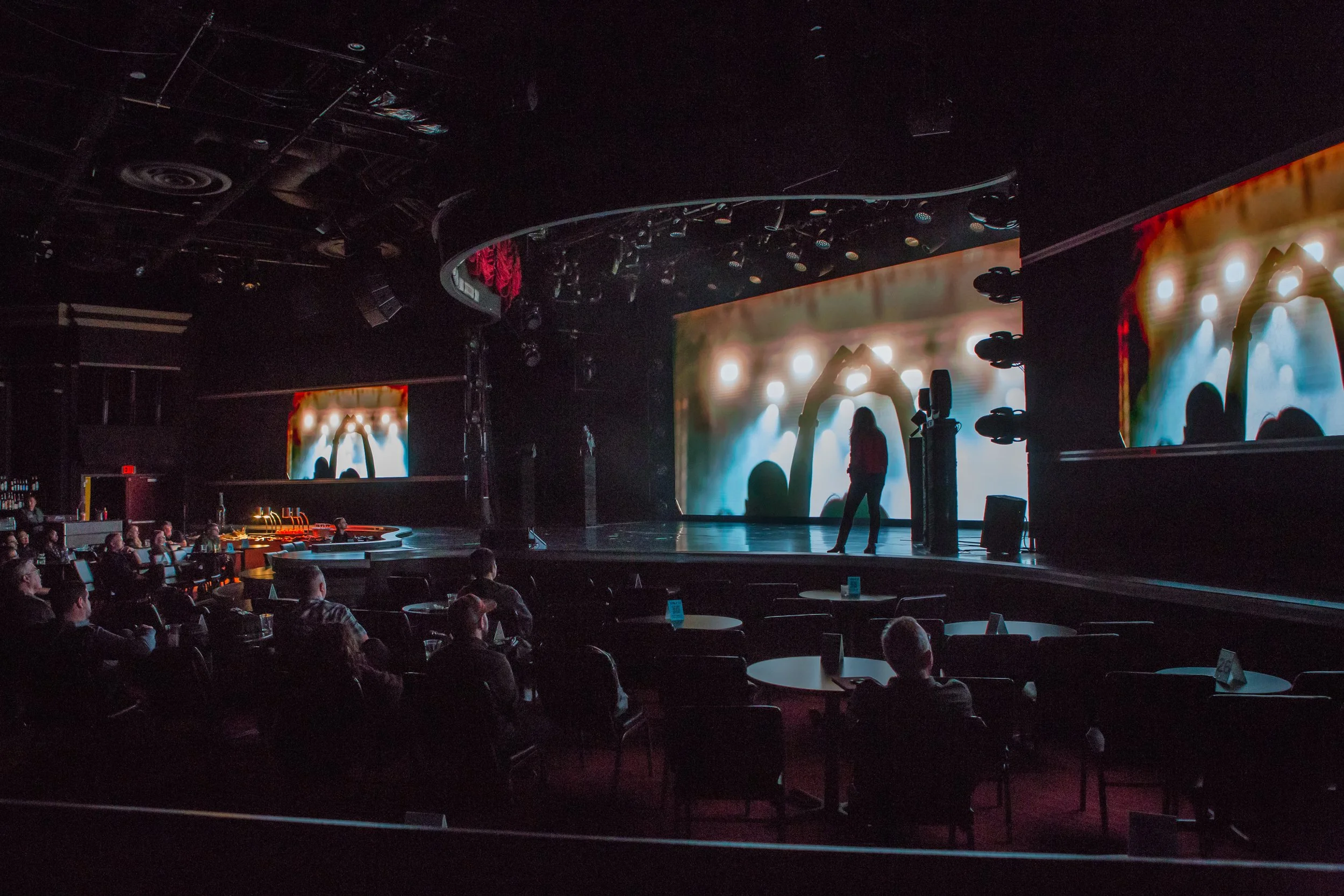 Dark theater with audience watching a stage performance, large screen displays colorful abstract visuals, a person stands on the stage, and stage lighting illuminates the scene.