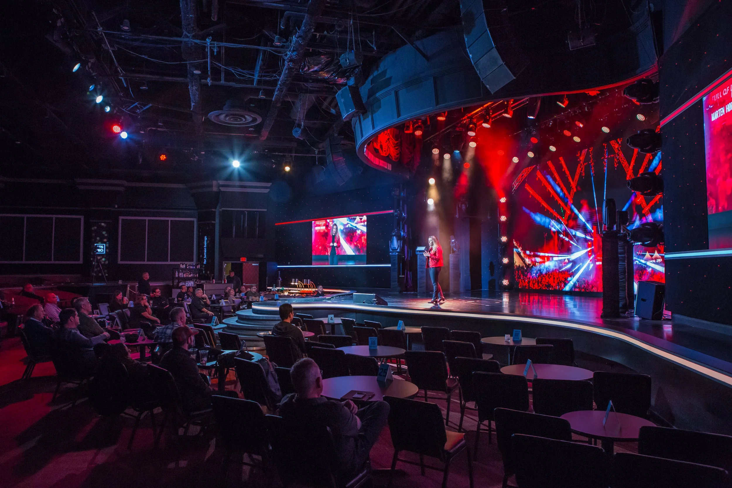 An indoor stage with bright lights, a large digital screen displaying a performer, and an audience seated in front.