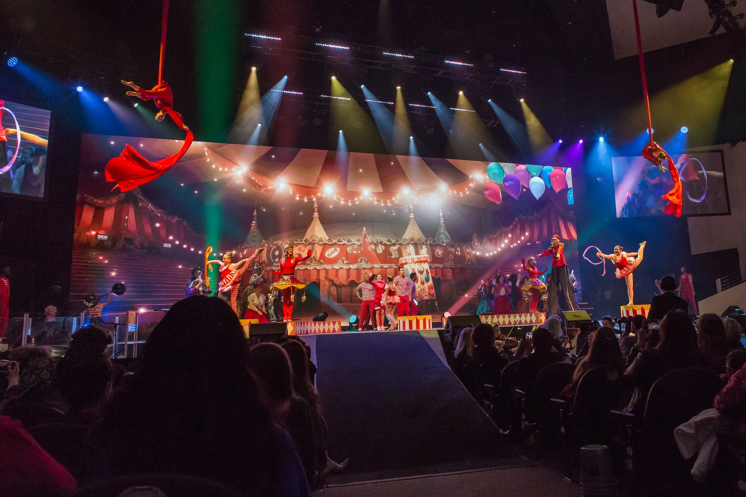 A lively circus performance on stage with acrobats, clowns, and performers in colorful costumes, featuring hanging red silks, bright lights, and a decorated backdrop with balloons and a striped tent design, while an audience watches.