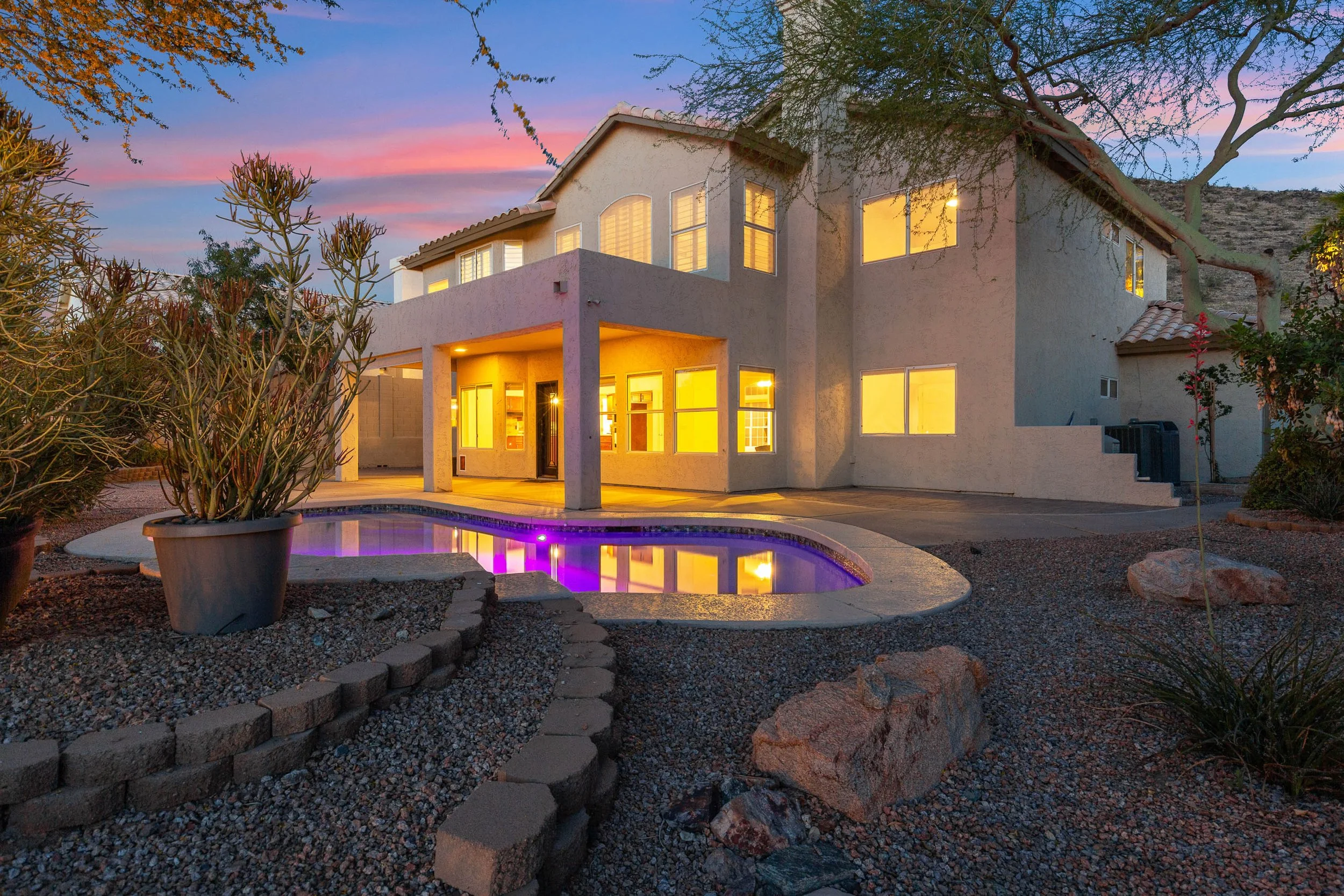 A modern two-story house with illuminated windows at dusk, a small purple-lit pool in the backyard, desert landscaping with rocks and potted plants, and a mountain hillside in the background.