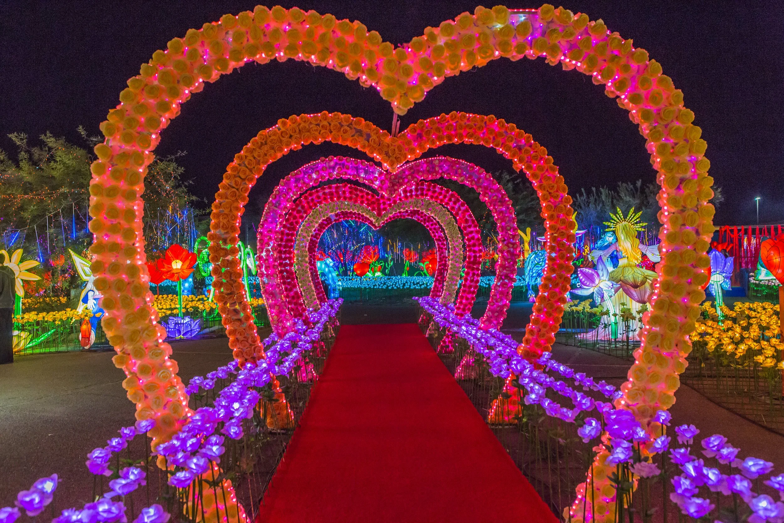 Colorful illuminated hearts made of flowers and lights form a tunnel at a nighttime outdoor event, with bright flower and fairy decorations in the background.