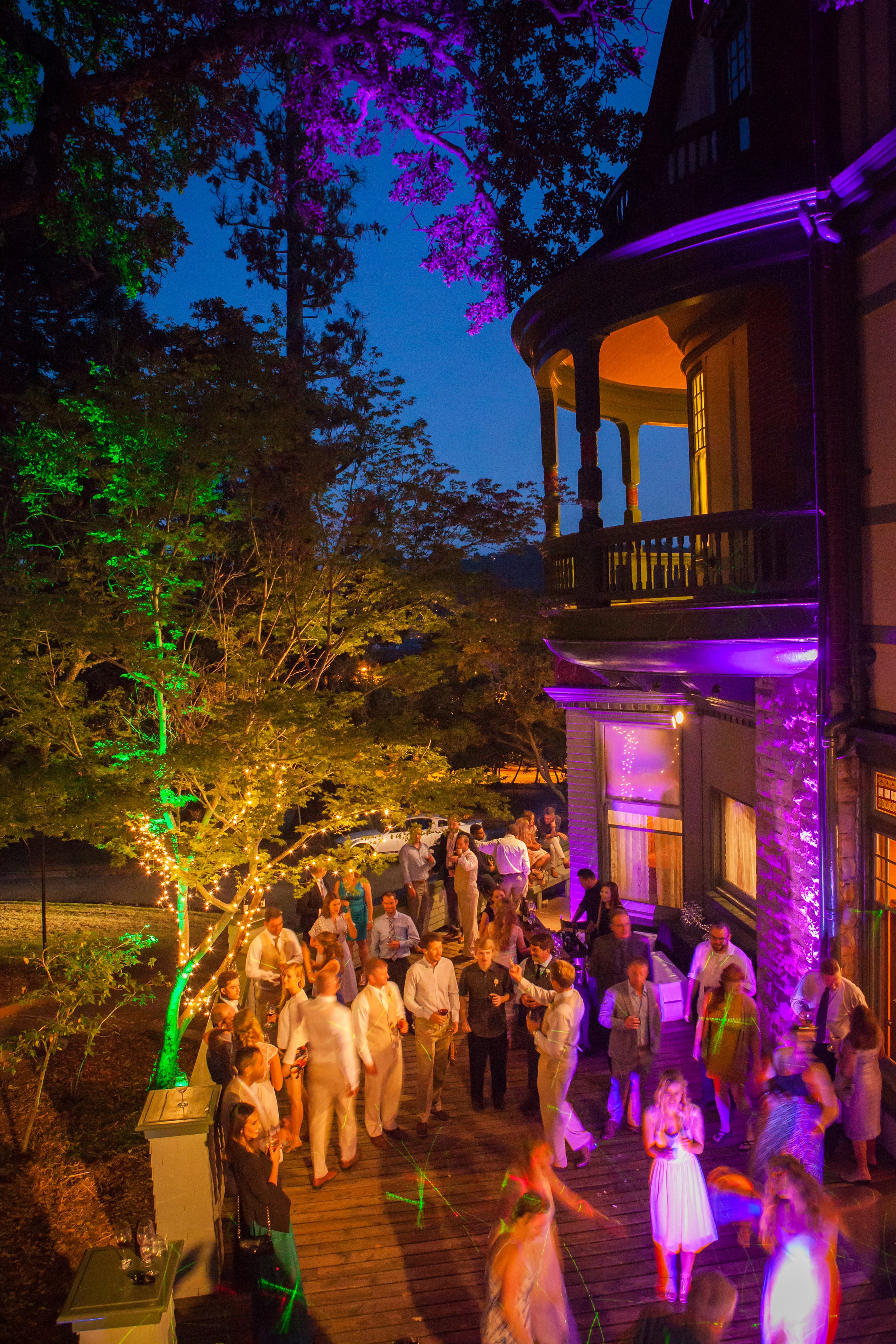 A lively outdoor party on a wooden deck at night, with people socializing, dancing, and drinking. The house has a balcony, and trees are decorated with colorful lights, creating a festive atmosphere.