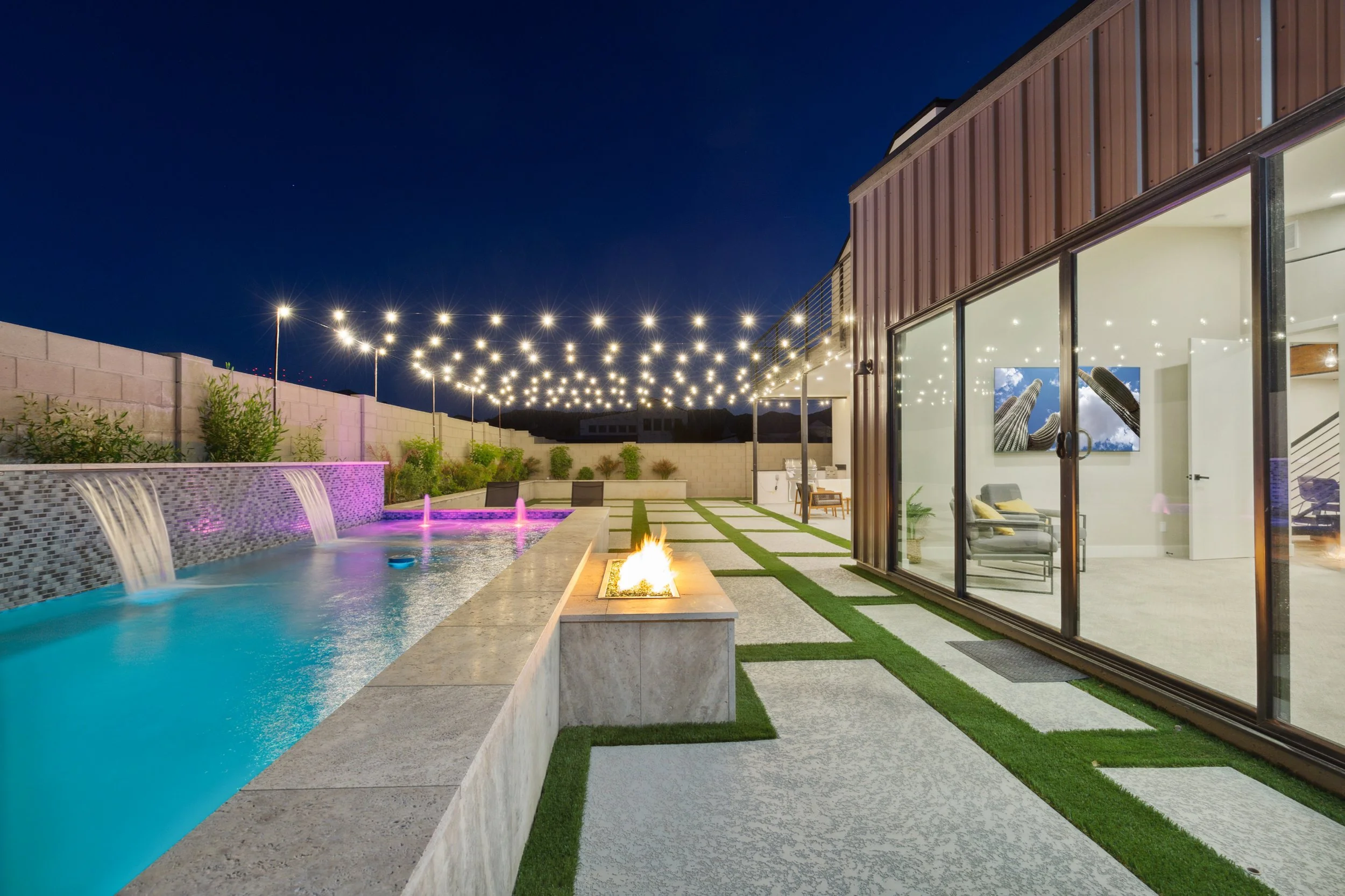 Nighttime backyard with a lit fire lounge area, pool with waterfalls and colored lighting, string lights overhead, and a modern house with large glass windows showing a living room inside.
