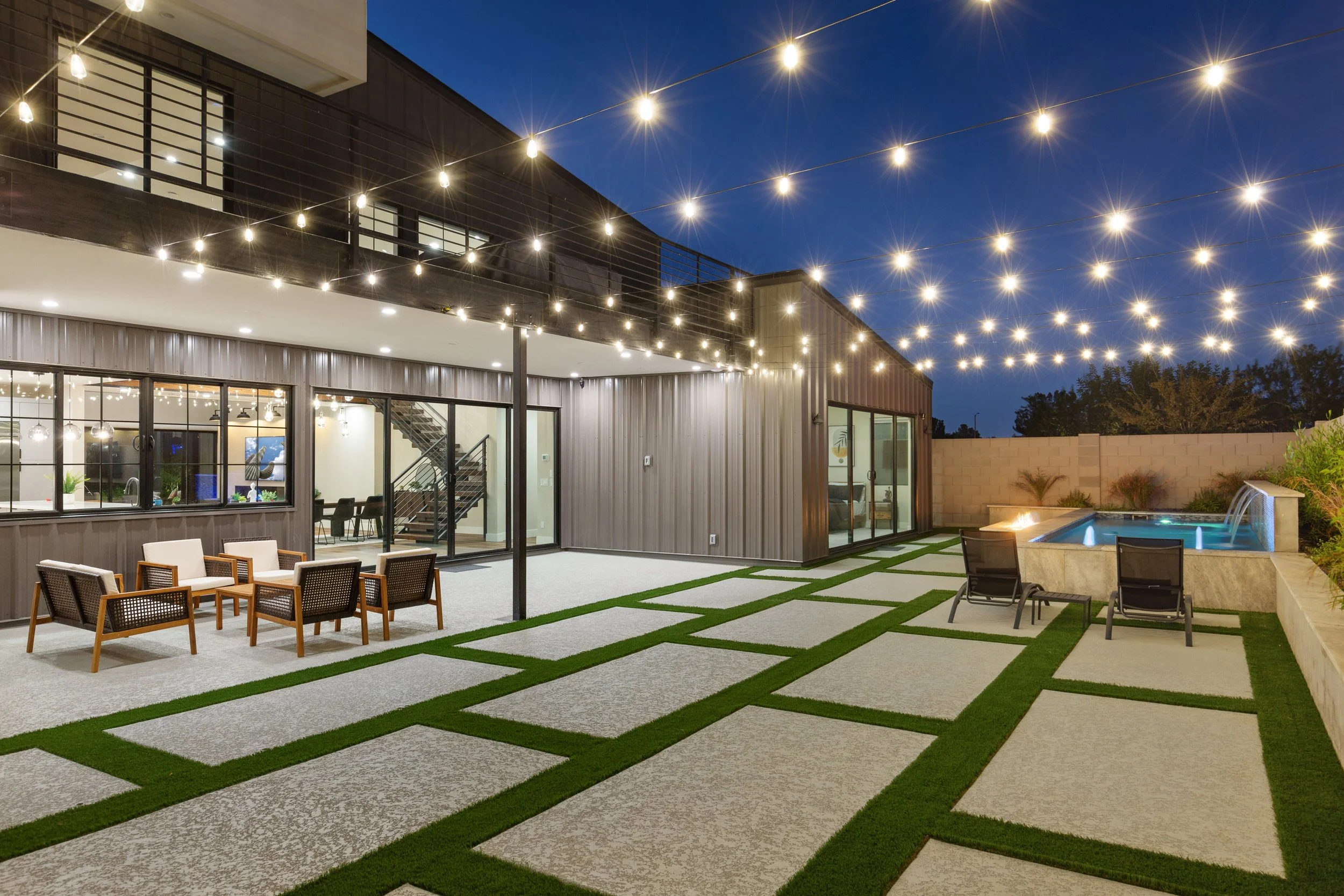 Modern backyard patio at night with string lights overhead, outdoor seating, a hot tub, and landscaping.