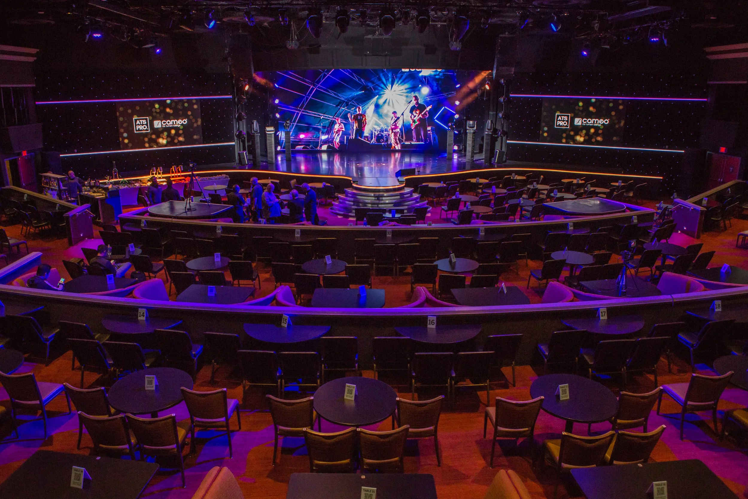 An empty theater or concert hall with a stage, where a band is performing with lighting effects, and seating arranged in front of the stage including tables and chairs.