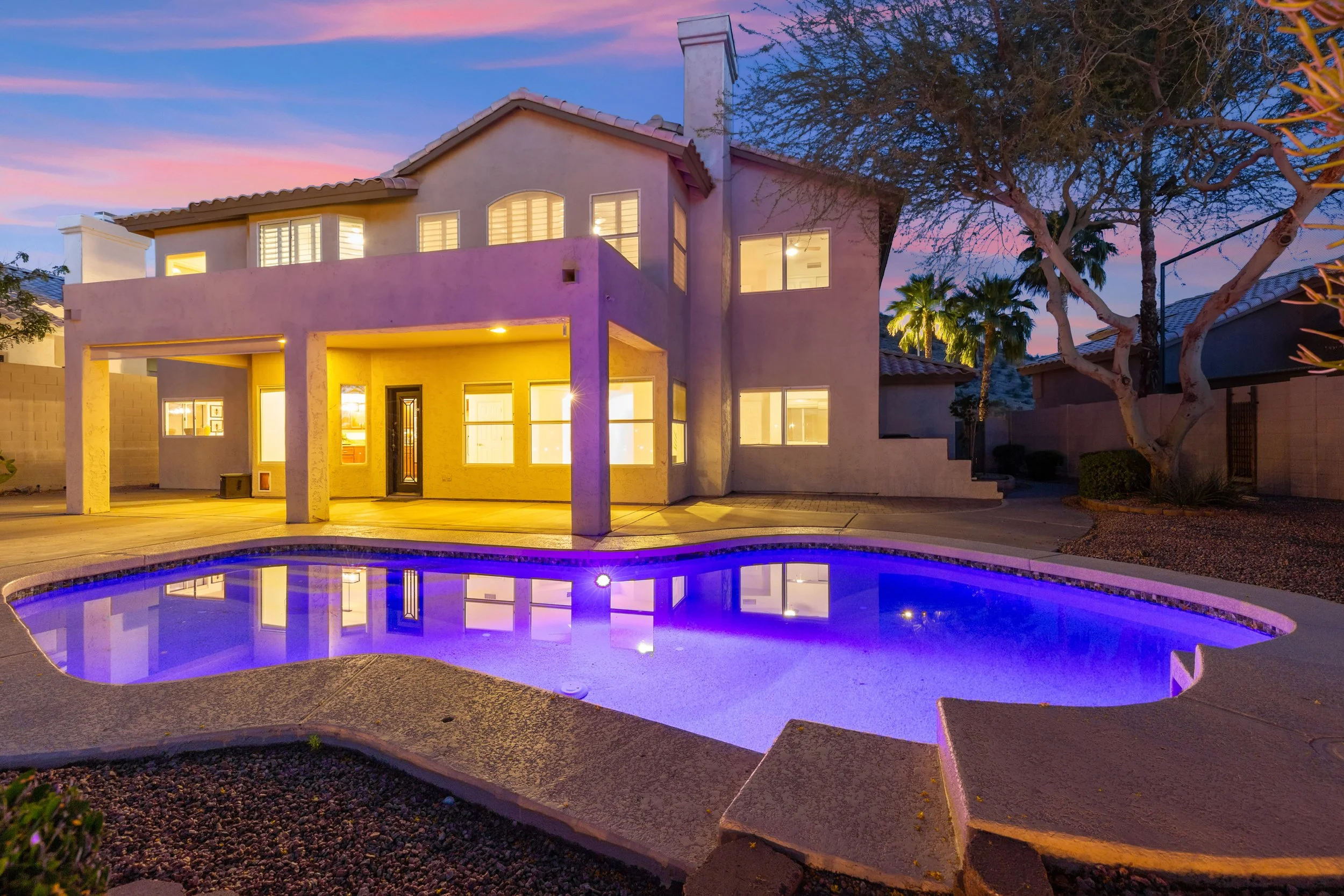 A two-story house with a backyard swimming pool during dusk, illuminated by interior and exterior lights.